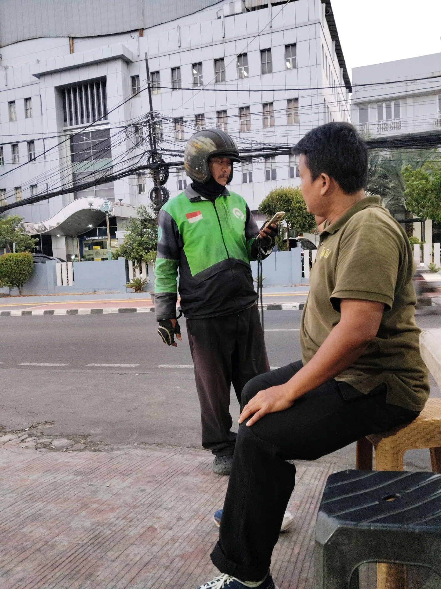 Two men are standing on a sidewalk next to a street in front of a modern building. The man on the left is wearing a green and black jacket with a helmet and holding a smartphone. The man on the right is wearing a green polo shirt and black pants and is sitting on a stool. The building in the background is a light-colored, multi-story structure with many windows and a prominent architectural feature at the entrance. Power lines are visible in front of the building. The street has white and yellow lines indicating traffic lanes. The sidewalk is made of concrete with textured lines. There are some trees and bushes on the side of the road.