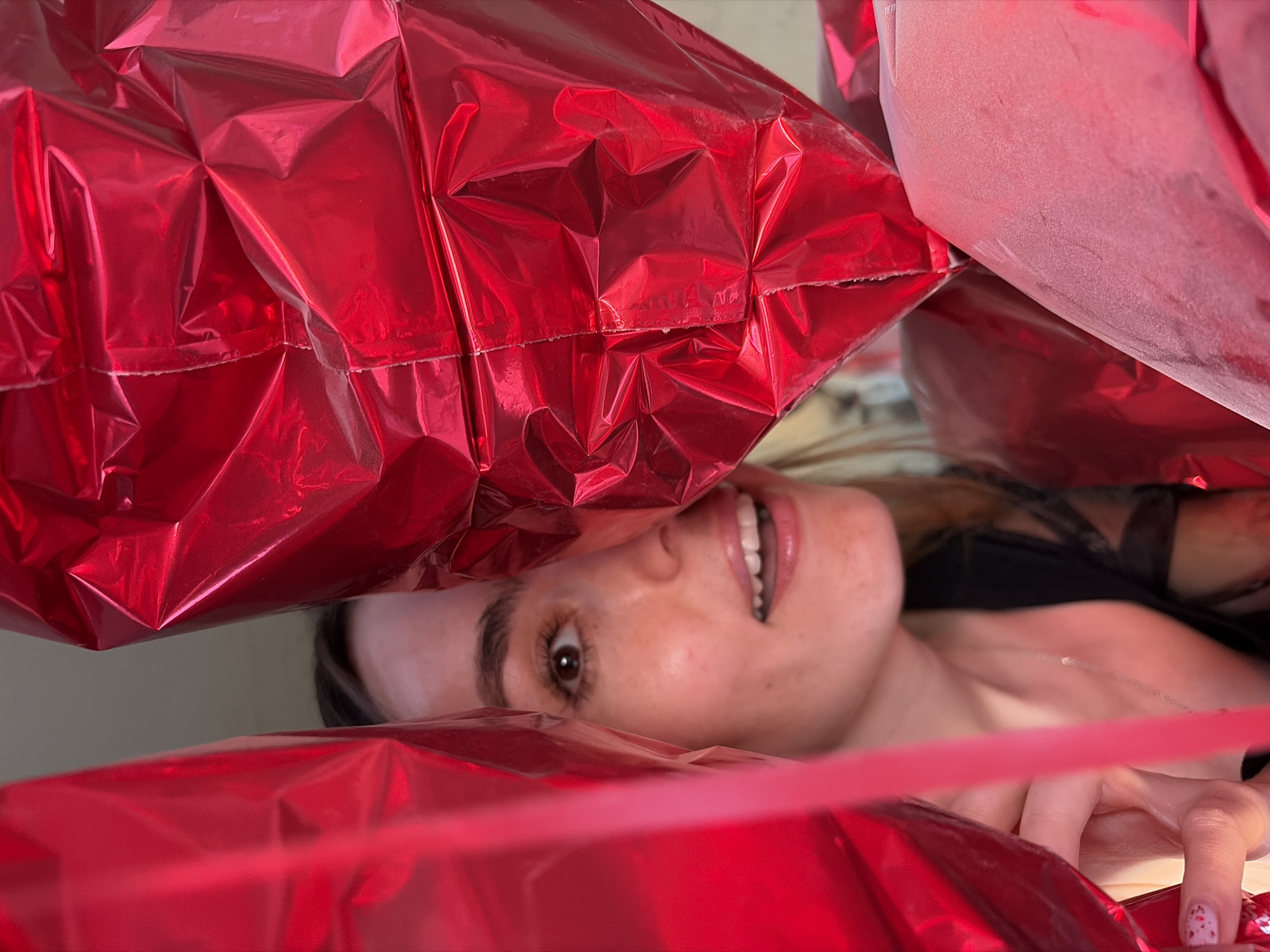 A person with fair skin, light brown hair, and brown eyes is partially obscured by red, crinkled, metallic balloons. The person's face is angled upward towards the camera, and a small portion of their chest and hand is visible. The background appears to be a plain, light-colored wall. There is no indication of the location in Izhevsk, Russia.