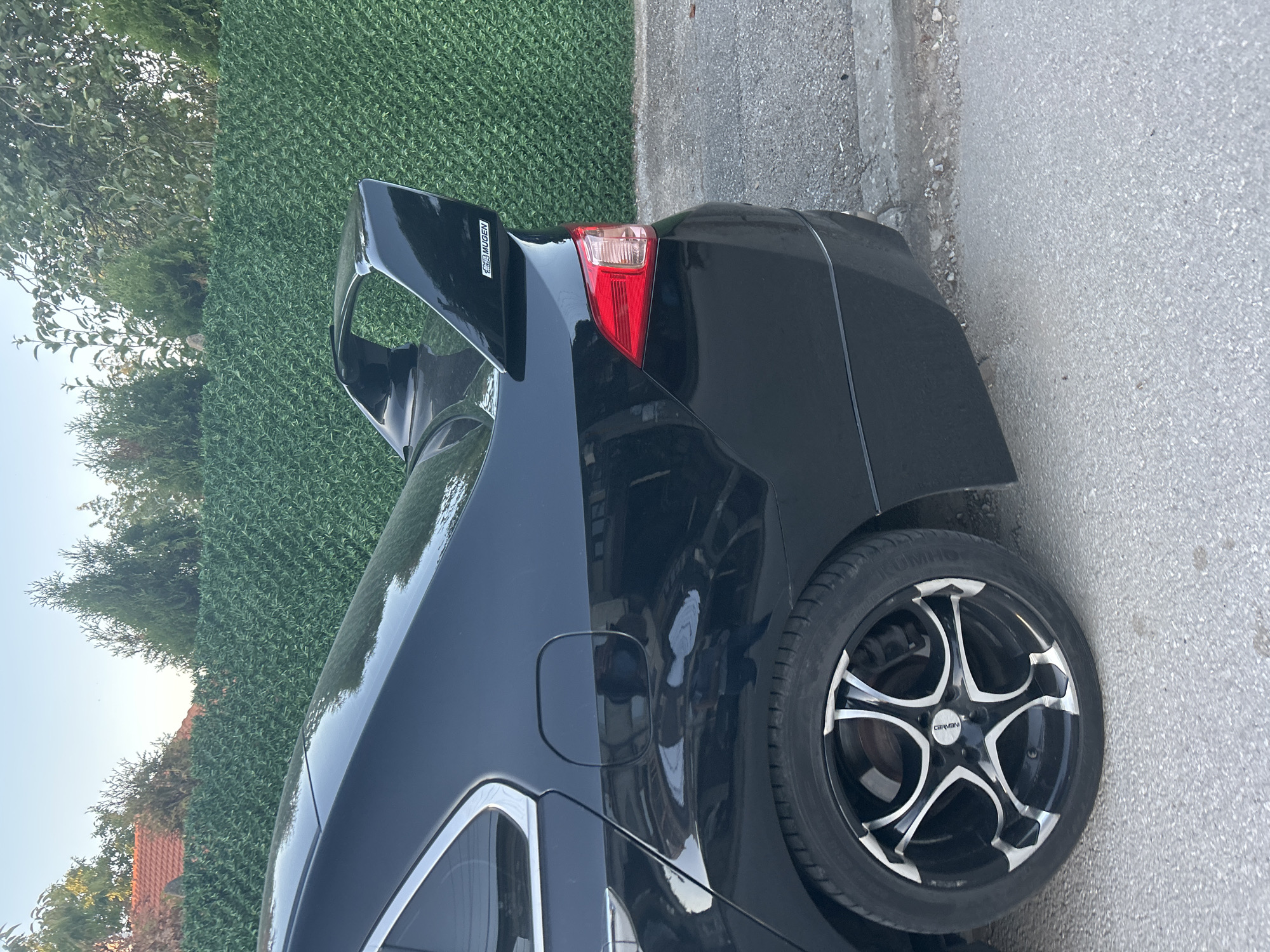 A black Honda sedan is parked on a paved road next to a green hedge. The car has a large rear spoiler and aftermarket rims. The spoiler has a "MUGEN" decal on it. The background shows trees and a residential building. The lighting suggests it is late afternoon or early evening.