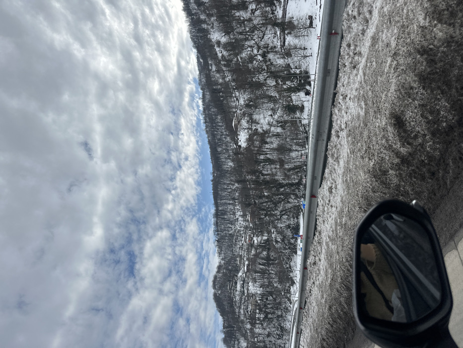 An outdoor, eye-level photograph captured from within a vehicle shows a snow-covered mountainous landscape in Ubisa, Georgia, under a partially cloudy sky. In the foreground, the side mirror of the vehicle is visible on the left, reflecting a partial view of an occupant wearing light-colored clothing. Adjacent to the vehicle and running parallel to the road is a snowbank composed of dirty, slushy snow. A grey metal guardrail separates this snowbank from a multi-lane highway, where blue and red markers are visible along the shoulder. Beyond the highway, a steep mountainside rises, densely covered with leafless deciduous trees and patches of snow. Several residential structures with dark roofs are scattered across the snow-dusted mountainside. The sky above is largely covered by stratocumulus clouds, with intermittent patches of clear blue sky visible. The overall scene suggests a winter day with recent snowfall.