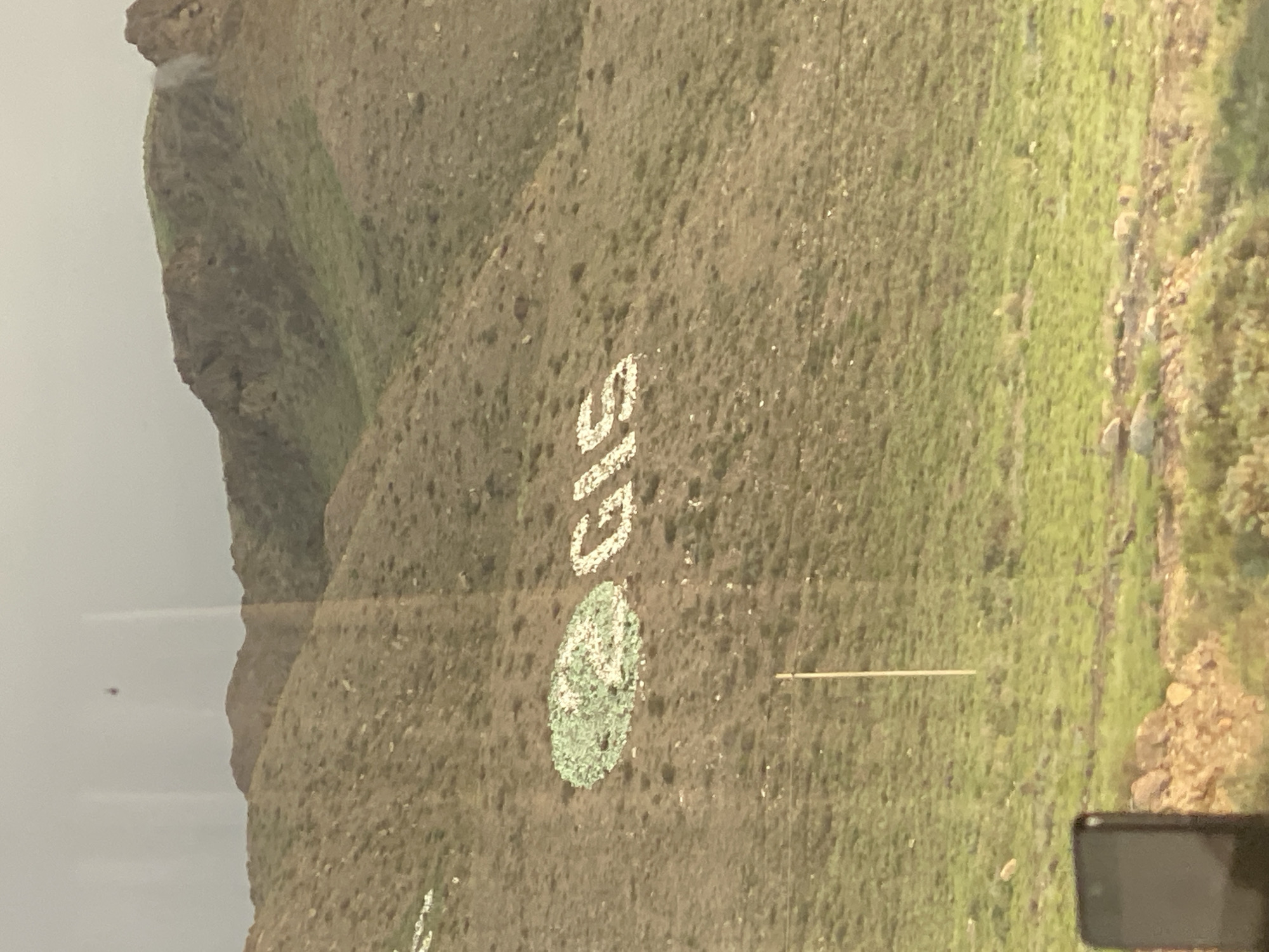 The image shows a hillside in the city of Kok-Moinok - 1, Kyrgyzstan. The hillside is covered with sparse vegetation and rocks. Large white letters spelling "GIS" are formed on the hillside using rocks. To the left of the letters, there is a circular formation of green and white rocks. A single utility pole stands vertically on the hillside below the letters. In the foreground, the top of a dark-colored smartphone is visible. The sky is overcast, and the overall lighting suggests a dull, possibly rainy day.