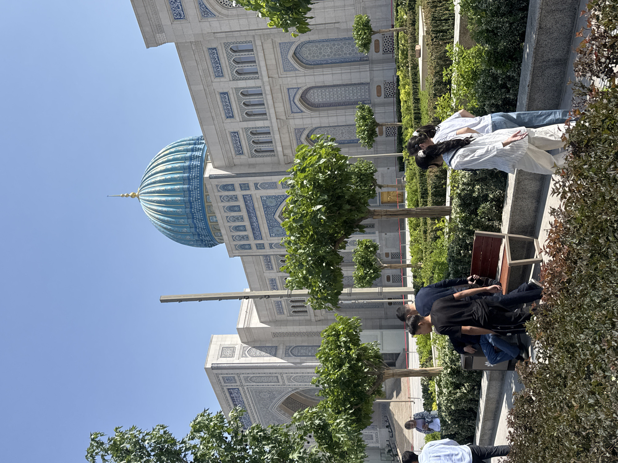The image shows the exterior of the Khast Imam complex in Tashkent, Uzbekistan, during daylight. The Hazrat Imam Mosque, characterized by a large, blue dome and white, intricately decorated walls, dominates the background.
In the foreground, there are people in various positions and activities. Some are sitting on benches, while others are standing or walking. The vegetation includes meticulously maintained trees and hedges, arranged along pathways and garden areas.
The sky is clear and blue, suggesting a sunny day. The overall scene presents a public gathering area within a prominent religious and cultural site.
