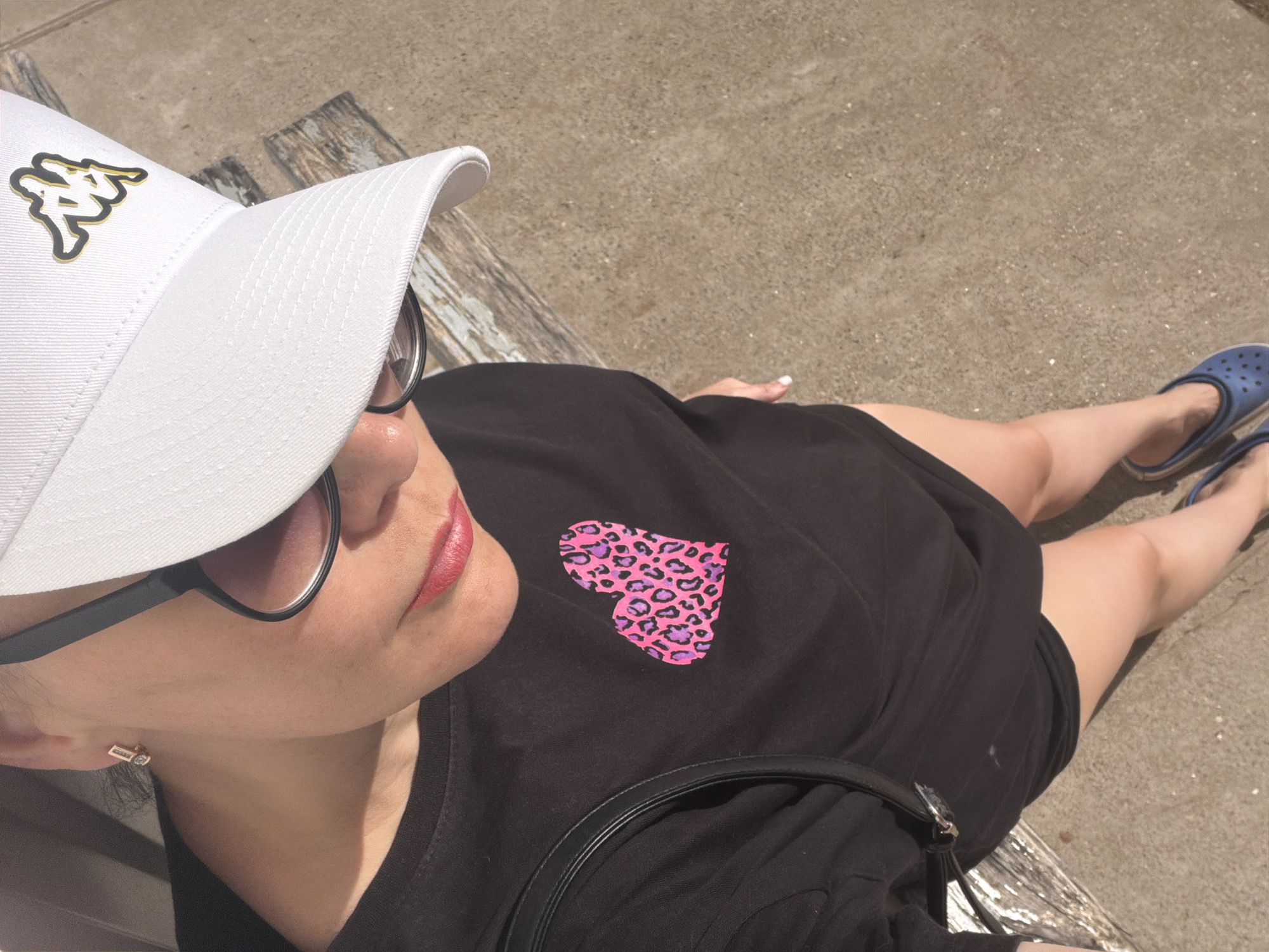 A person sits on a bench. The person is wearing a white baseball cap with a gold logo, black-framed glasses, and a black t-shirt with a pink leopard print heart. The person is also wearing pink lipstick and has short brown hair. The person is wearing blue Crocs. The location of this image cannot be identified.