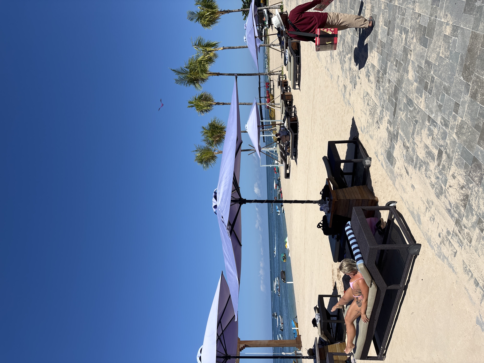 The image depicts a sunny beach scene in Nusa Dua, Indonesia, with a clear blue sky overhead. Palm trees line the beach, and several white sun umbrellas provide shade for beachgoers. A woman in a pink bathing suit reclines on a dark-colored lounge chair. In the background, there are multiple boats in the water. A man in a red long-sleeve shirt, khaki pants, and a white hat stands near a brick walkway. A red kite is visible in the sky above the palm trees.