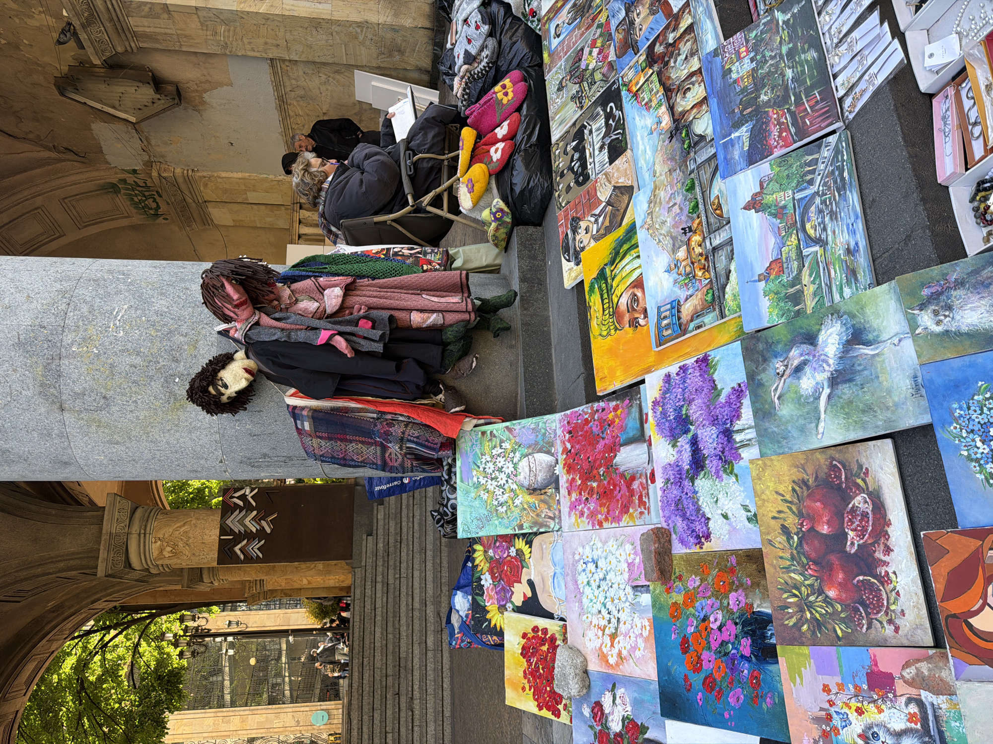 The image depicts an outdoor art market in Tbilisi, Georgia. A vendor sits on a folding chair behind an array of paintings displayed on the ground. The paintings vary in subject matter, including landscapes, portraits, still lifes with flowers and pomegranates, and abstract compositions. Beside the vendor are bags containing items like slippers and clothing. A dressed doll lies against a stone column that forms part of an arched structure. To the left, a street scene is visible through the archway.