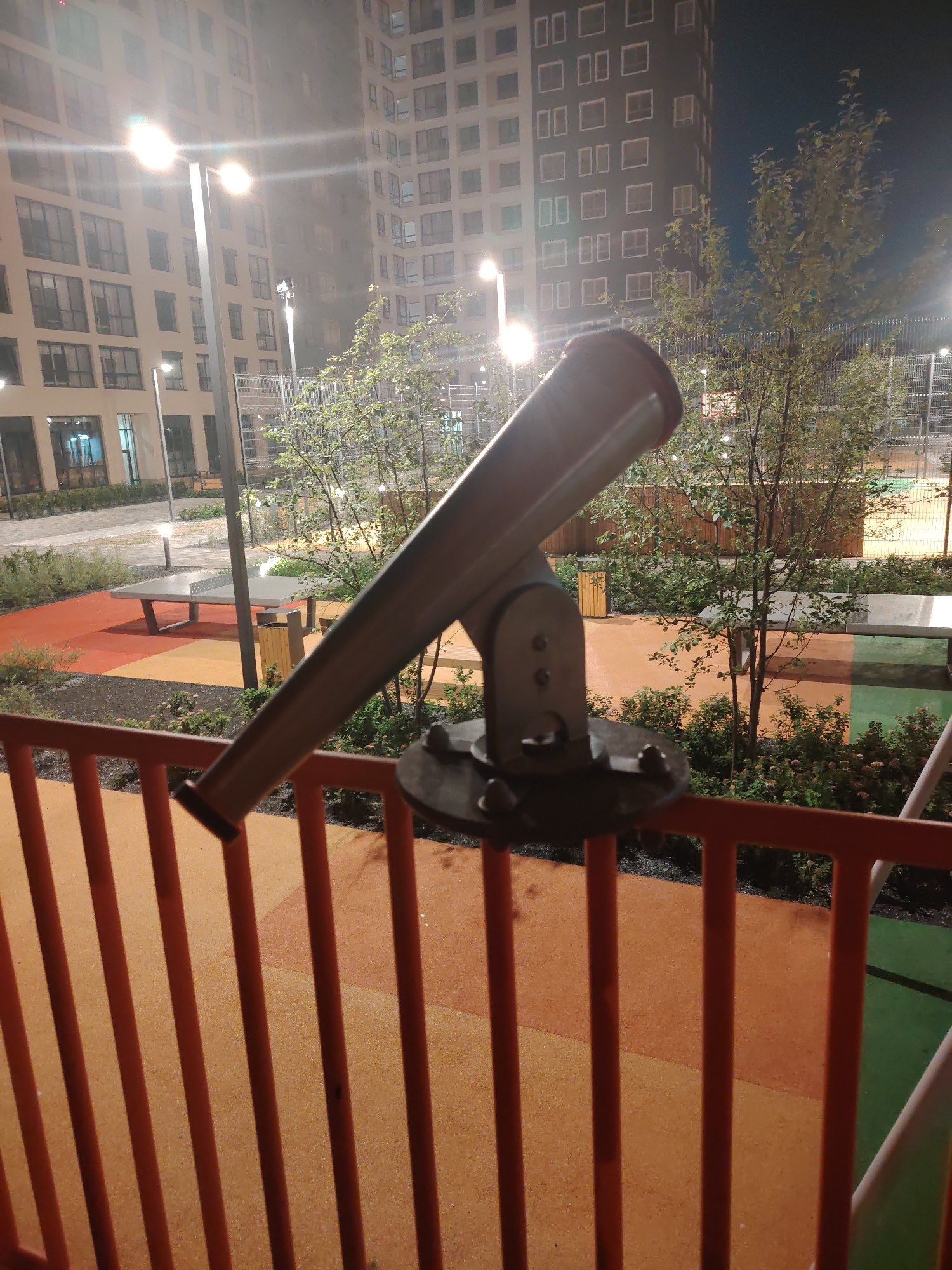 The image captures a nighttime view of an outdoor recreational area with a telescope mounted on a railing in the foreground. Behind the telescope, modern apartment buildings with numerous illuminated windows rise into the dark sky. The ground level features a colorful surface with distinct sections of orange and yellow, a ping pong table, and a trash receptacle. Trees and dense shrubs are visible in the midground, and a fence encloses the area. The scene is illuminated by artificial lighting, including streetlights and lights from the buildings. The image does not contain any individuals or explicit actions. The presence of apartment buildings and recreational facilities suggests an urban or suburban environment, possibly a residential courtyard or park. Based on the style of architecture and the presence of specific street furniture or design elements, this scene is likely located in the city of Yekaterinburg, Russia.