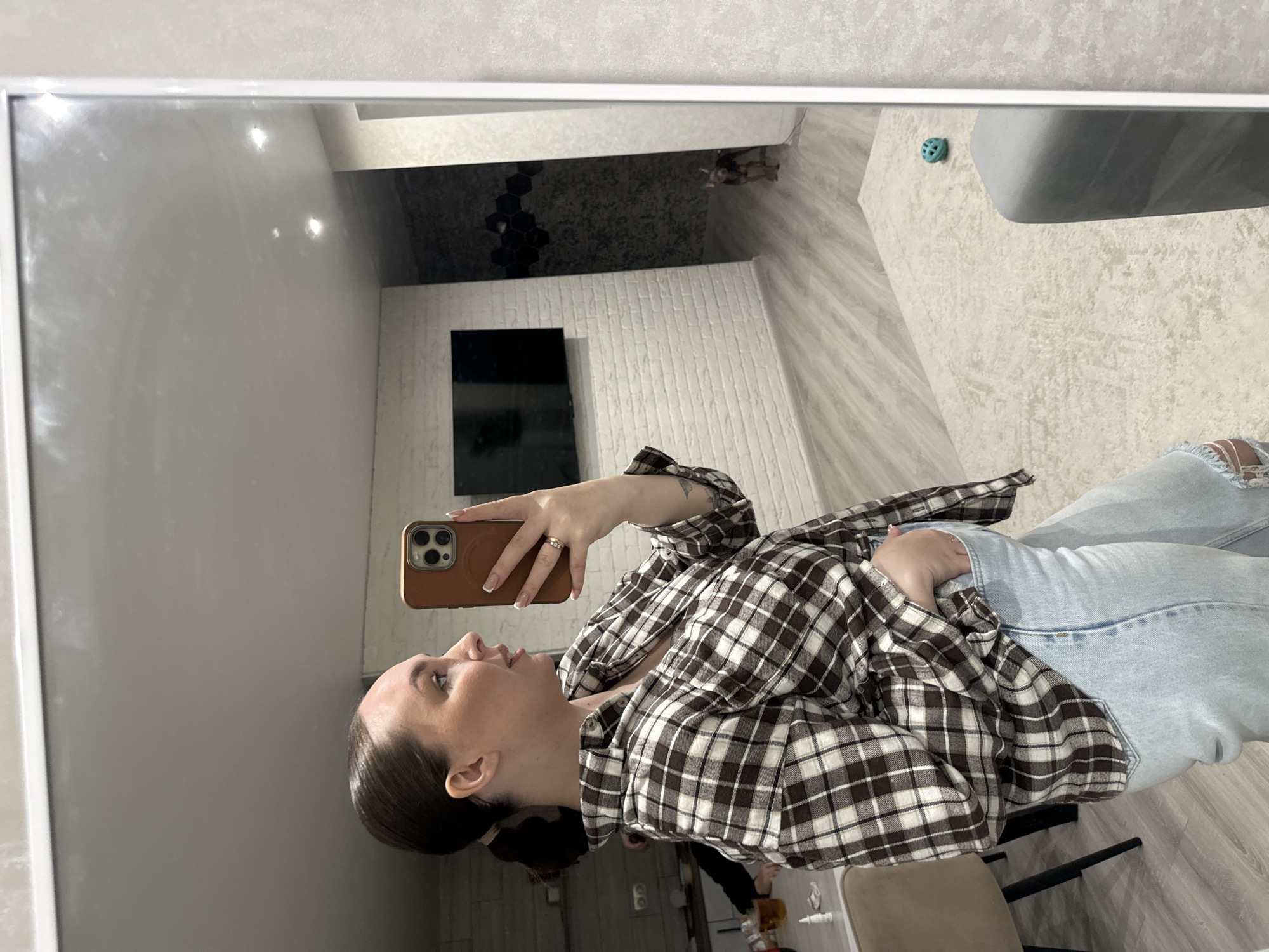 A mirror selfie taken indoors shows a young woman in what appears to be a modern living room or bedroom in Chaikovsky, Russia. The woman, who appears to be in her 20s or early 30s, is positioned on the right side of the frame. She has dark hair styled neatly, likely pulled back. She is wearing a casual outfit consisting of a brown and white plaid flannel shirt and light blue ripped jeans. A ring is visible on her left hand's ring finger. She holds a smartphone with a brown case in her right hand, pointed towards the mirror to capture her reflection. Her expression is neutral, and she appears to be looking at the phone screen.
The mirror reflects the room behind her, which is well-lit, suggesting daytime or bright artificial lighting. The floor is light wood laminate, partially covered by a large, light beige or white textured rug. A grey sofa or armchair is visible on the rug, and a small, blue spherical toy rests nearby. In the background, a wall with a light-colored brick texture features a mounted dark rectangular object, possibly a television or screen. Above this, a darker wall section with hexagonal textured tiles is visible. A small, light-colored animal, likely a cat, can be faintly seen near the corner where the dark and light walls meet, on the floor. A brown chair or stool leg is partially visible in the bottom right foreground reflection. No text is visible in the image.
