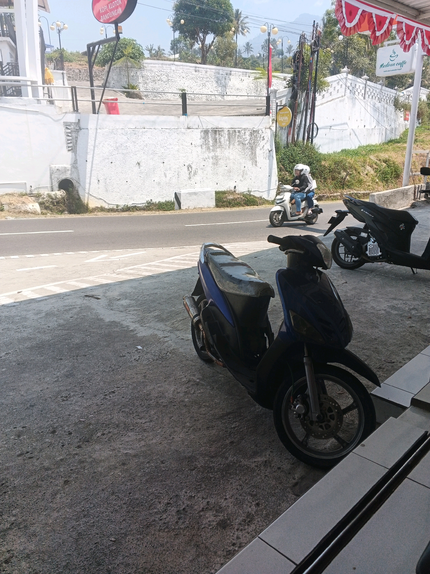 The image captures a roadside scene in Pakopen, Indonesia, focusing on two parked scooters and a section of a road with passing traffic. In the foreground, a dark blue scooter with a textured seat is parked on a concrete surface. To its right, a black scooter is also parked. The road, marked with white lines, is visible behind the scooters, with a white vehicle and a scooter carrying two passengers traveling away from the viewer. In the background, a white stone wall with decorative patterns lines the roadside, leading up a slope. A sign with "Medan Caffe" and a coffee cup logo is mounted on a pole near the wall. Further in the background, trees and buildings are visible under a bright sky. Red and white decorative flags are strung across the top right of the frame. The overall lighting suggests it is daytime with a clear sky.