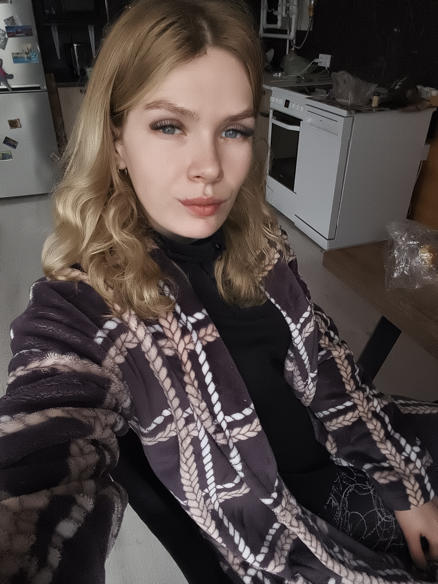A young woman with blonde, wavy hair is the subject of this indoor portrait. She wears a black turtleneck sweater under a brown robe with a white rope pattern. The woman is seated and faces the camera directly, with a slight smile. Her blue eyes are accentuated by dark eyelashes.
The setting appears to be a kitchen in a residential area. A white refrigerator with magnets is visible in the background to the left, while a white stove/oven unit stands to the right. A wooden table is partially visible in the right background. The location is potentially in Русский Вожой, Russia, based on the prompt.