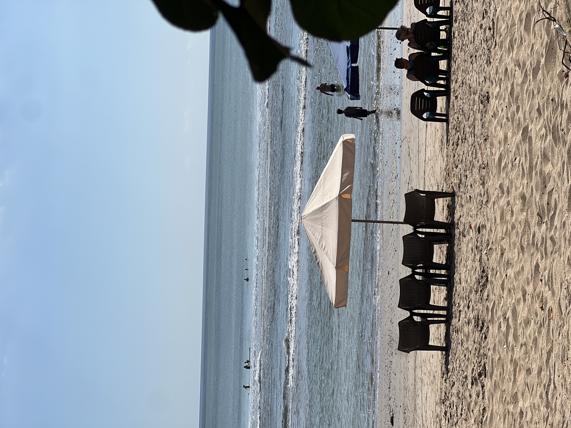 A wide-angle, eye-level shot captures a serene beach scene in Kuta, Indonesia. The foreground is dominated by textured sand, leading to a calm ocean with gentle waves lapping the shore. Several beach chairs are scattered across the sand, some occupied by individuals relaxing. A prominent beige umbrella provides shade on the beach. In the middle ground, the ocean is populated by a few surfers and swimmers, some with surfboards. The horizon is a clear, pale blue sky, with no clouds visible. The overall ambiance is tranquil and inviting, suggesting a typical day at a popular Indonesian beach destination.