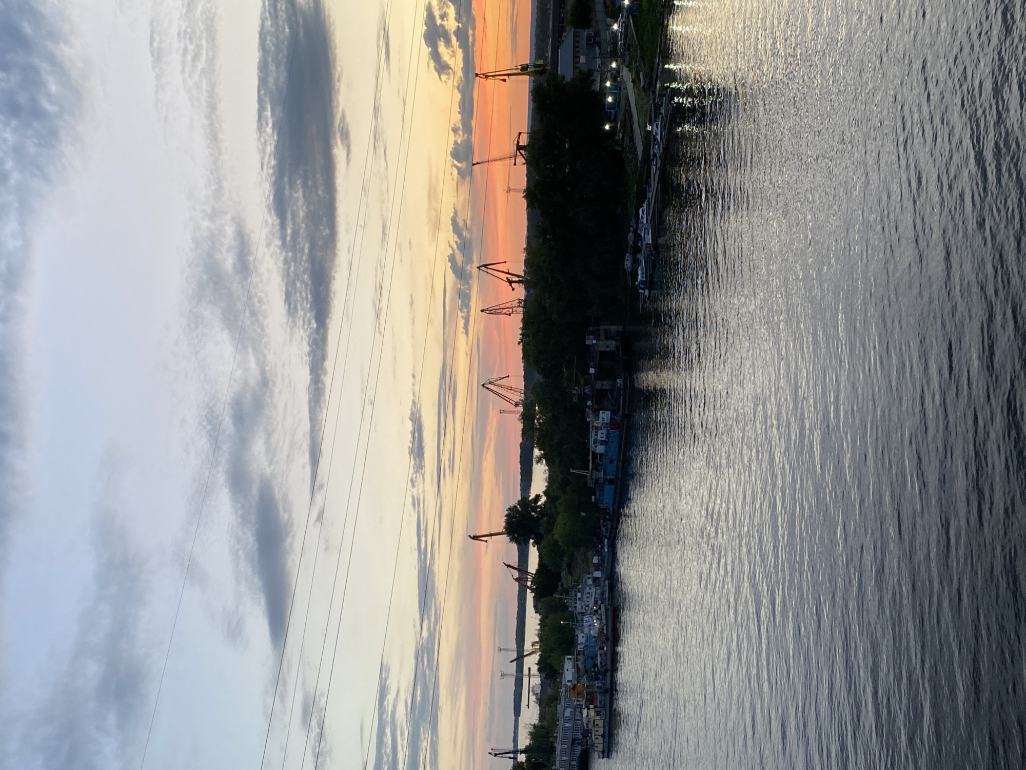 The image depicts a waterfront scene at dusk in Novokuybyshevsk, Russia. The sky is a canvas of soft colors, transitioning from pale blue and grey clouds to warm hues of orange and pink near the horizon, indicating sunset. The foreground is dominated by a body of water, with gentle ripples catching the fading light, creating a shimmering effect.
Along the bank, a line of trees separates the water from an industrial area. Several large cranes, characteristic of a port or industrial yard, are visible against the colorful sky. These cranes suggest ongoing or recent activity, likely related to cargo handling or construction. Various vessels, including boats and barges, are docked along the waterfront, adding to the sense of an active port.
Overhead power lines stretch across the sky, a common sight in urban and industrial landscapes, and they create diagonal lines that add a geometric element to the composition. The lighting suggests it is late evening, with artificial lights from the dock area beginning to illuminate the scene. The overall mood is peaceful and contemplative, with the natural beauty of the sunset contrasting with the industrial elements of the location. There is no visible text in the image.