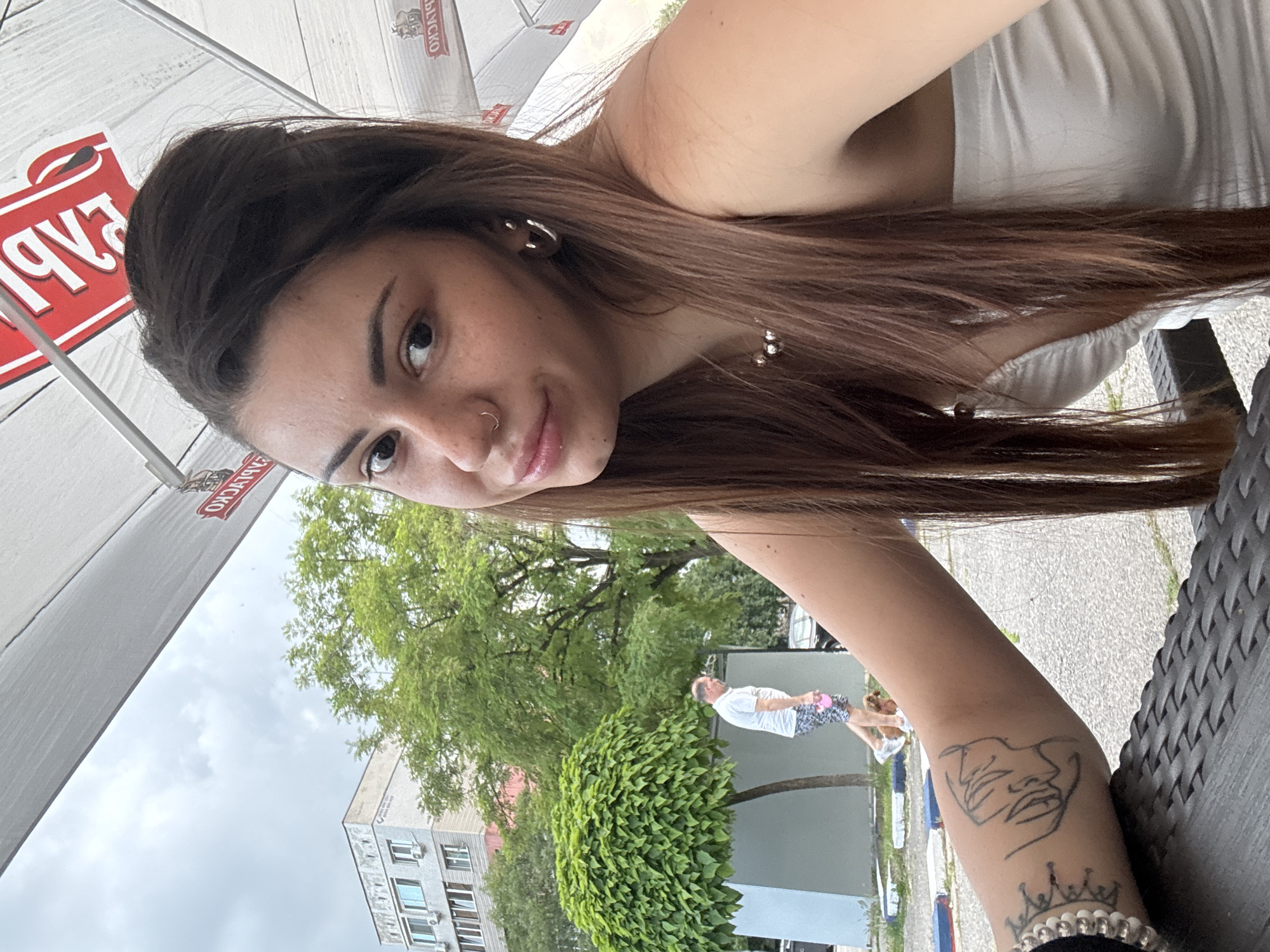 A young woman is depicted in a close-up selfie, seated outdoors under a white awning. She has long, dark brown hair, dark eyes, and visible freckles on her face. She wears a small hoop nose ring on her left nostril and a silver hoop earring in her right ear. A silver beaded necklace is visible around her neck, and she is wearing a white strapless top. Her left forearm, resting on a dark woven-patterned table, displays two tattoos: a small crown-like design near her wrist and an abstract linear drawing resembling a face further up her arm. A silver beaded bracelet is also on her left wrist.
Above her, the white awning features inverted red logos, one clearly identifying "ШУМЕНСКО" (Shumensko), a Bulgarian beer brand, and another less distinct logo, suggesting an outdoor cafe or restaurant setting. The sky beyond the awning is overcast. In the background, an urban environment is visible, including a multi-story building with multiple windows and air conditioning units. Several green trees, one with a distinctive rounded canopy, are present. A man wearing a white polo shirt and patterned shorts is walking in the mid-ground, holding a pink leash with a small brown dog beside him. A dark vehicle is partially visible behind them. The ground is paved, and blue and red painted curb markings are discernable.
Based on the "ШУМЕНСКО" beer logo, the location is identified as Sofia, Bulgaria, within an outdoor seating area of an establishment in a city setting.