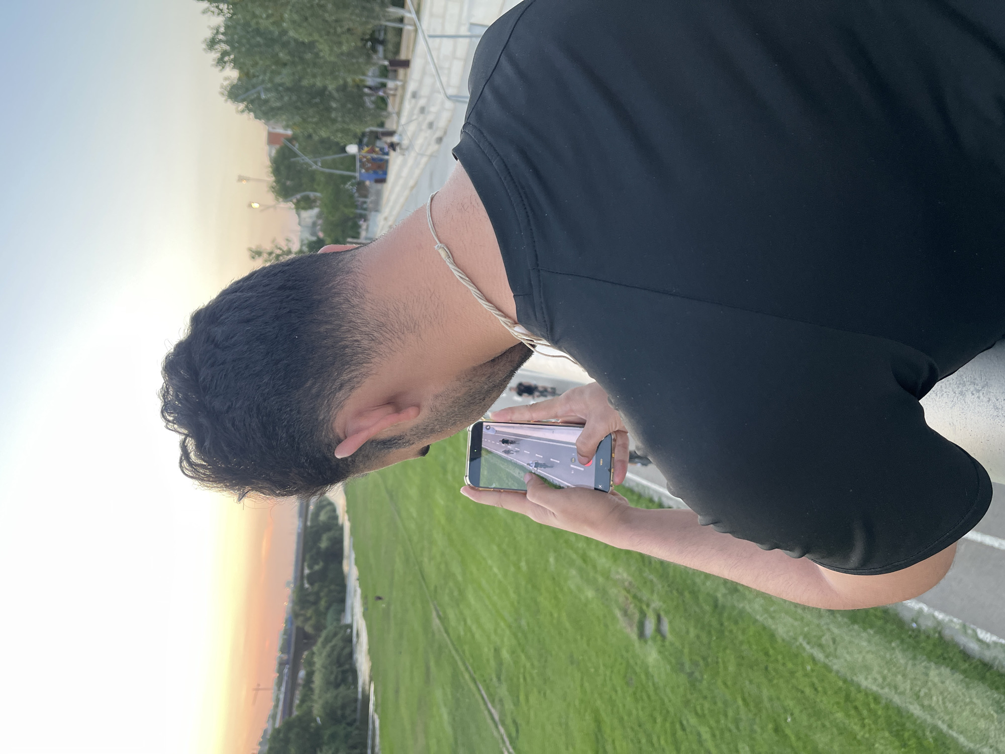 The image shows a person from behind, holding a smartphone in their hands and looking at its screen. The person is wearing a black t-shirt and a braided necklace. The smartphone displays a video of cyclists on a road.
The background reveals a park-like setting with a river or body of water on the left, and grassy slopes. In the distance, a bridge is visible spanning across the water, and beyond that, a city skyline with buildings and trees. The sky is a soft gradient of orange and blue, suggesting it is either sunrise or sunset. The overall environment appears to be a public space, possibly a park or promenade, in Santa Coloma de Gramenet, Spain, given the presence of a river, urban development, and distinct vegetation characteristic of the area. There are other people visible in the distance, some appearing to be cycling. The image is taken from a slightly elevated position, looking down towards the river and the park.