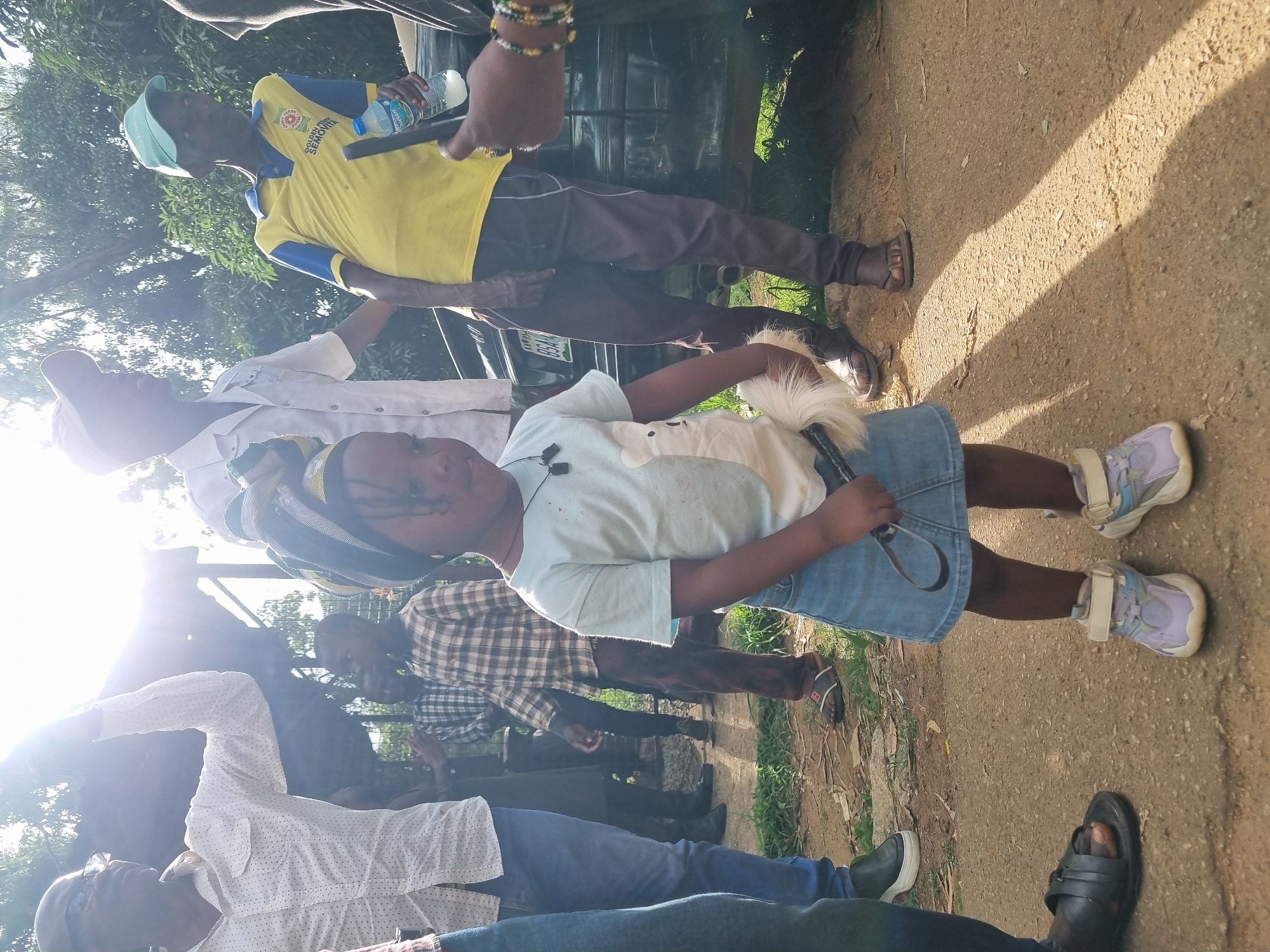 In Jos, Nigeria, a group of people are gathered outdoors. The scene is brightly lit, suggesting it is daytime, possibly late morning or early afternoon, with sunlight casting shadows on the sandy ground. The weather appears to be warm and dry.
The main subjects include several adults and a young girl. One adult, wearing a yellow and blue polo shirt with some text, is holding a bottle of water and a phone. Another adult, in a white patterned shirt, is visible in the foreground, wearing glasses. The young girl, in the center of the frame, is wearing a light blue t-shirt and a denim skirt, with purple and white sneakers on her feet. She is holding a leash attached to what appears to be a white dog or toy. Other individuals are partially visible in the background, some wearing plaid shirts.
The group seems to be socializing or perhaps preparing for an activity. There are cars in the background, indicating a roadside or parking area. The overall atmosphere seems casual and relaxed. The visible text on the yellow shirt reads "YOUTHS SEMO..." and also includes a logo with text that is difficult to decipher. A license plate on a car in the background shows "FGN YS8".