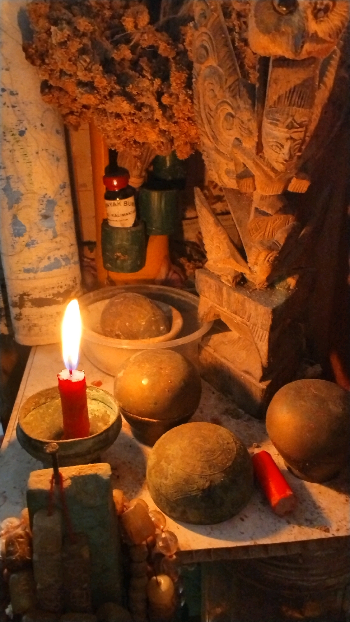 The image depicts an altar with various objects, including a lit red candle in a brass holder, several round, stone-like objects, a small bottle with a red cap and a label, a dried floral arrangement, and a carved wooden figure. A small, red cylindrical object is also visible near the spherical objects. The background includes a textured, light-colored surface and part of what appears to be a carved wooden structure. The lighting is dim and warm, suggesting an indoor setting, possibly at night or in a dimly lit room. The overall impression is one of a ritualistic or spiritual arrangement. The location is identified as Penompo, Indonesia.