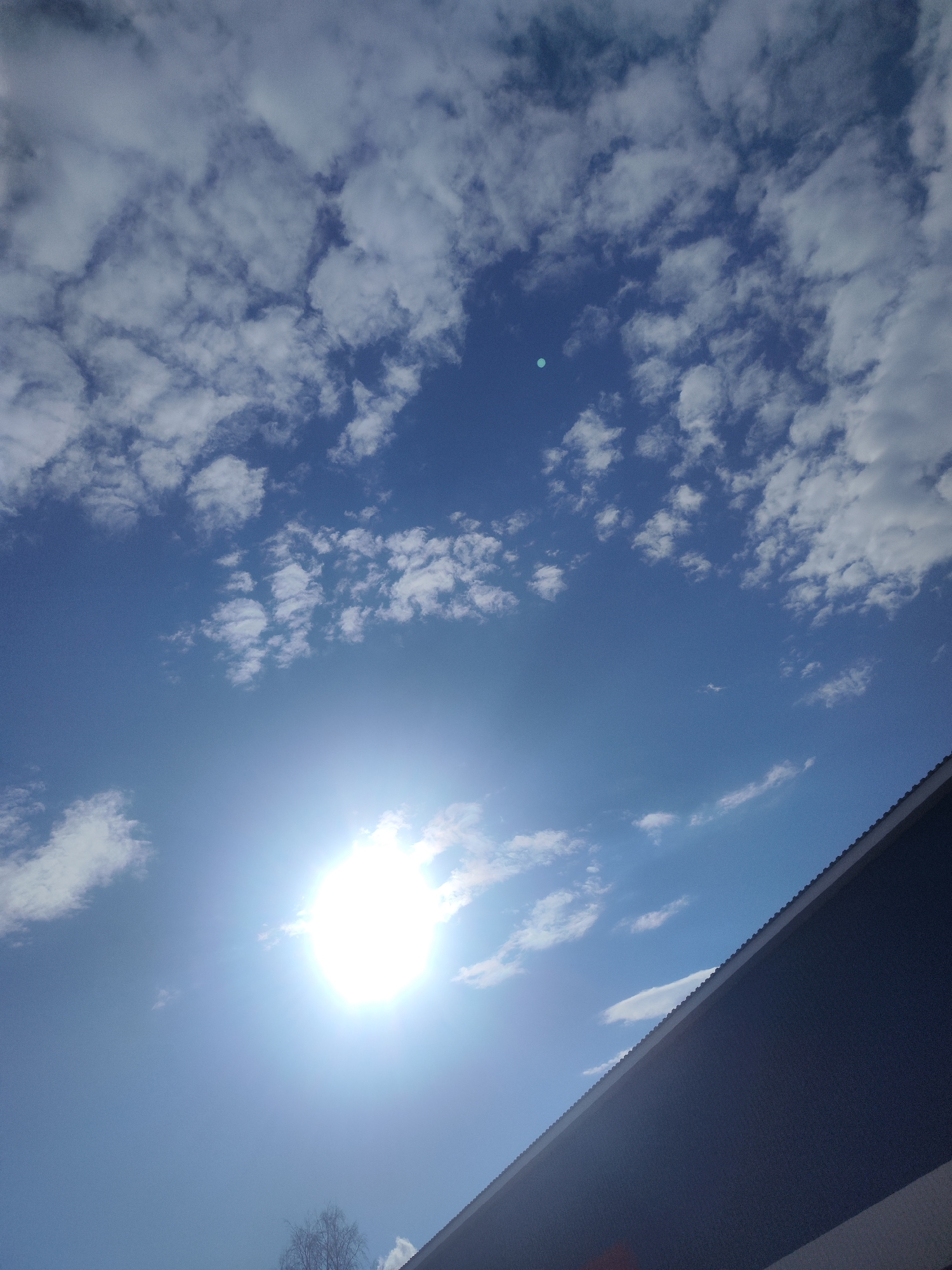 The media file presents an outdoor, upward-facing view of a bright blue sky filled with scattered white cumulus clouds. The sun, a brilliant and overexposed orb, is prominently located in the lower-left quadrant, acting as a strong light source and creating a distinct lens flare effect. A bright green circular artifact is visible in the upper-middle section of the sky, likely a photographic anomaly from the direct sunlight.
In the lower-right portion of the image, the dark, angled roofline of a building is visible, cutting diagonally across the frame. The roof appears to be made of a dark blue or grey material, possibly corrugated, and is either in shadow or dark in tone. In the very bottom-left corner, a few bare tree branches are subtly visible against the bright sky, suggesting dormant deciduous trees.
The scene depicts a clear, sunny day, likely around midday given the intensity of the sun. The weather appears fair with broken cloud cover. There are no people or explicit activities taking place. The setting is an outdoor area in Chaikovsky, Russia. No visible text is present.