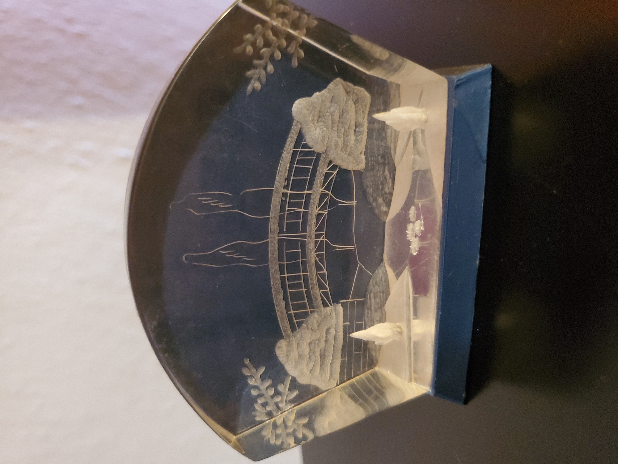 A clear, fan-shaped acrylic paperweight is pictured, showcasing an etched scene of a bridge with two figures walking across it. The bridge is arched, with a detailed railing. Two stylized trees flank the scene, and foliage details are etched on either side of the paperweight. At the base of the scene, a small body of water is visible, with two small white floral-like elements. The paperweight rests on a dark blue rectangular base, which in turn sits on a dark, reflective surface. The background is a plain, lightly textured off-white wall. There are no people present in the image, and the object is static. This image is not from Konstantinovka, Moldova.