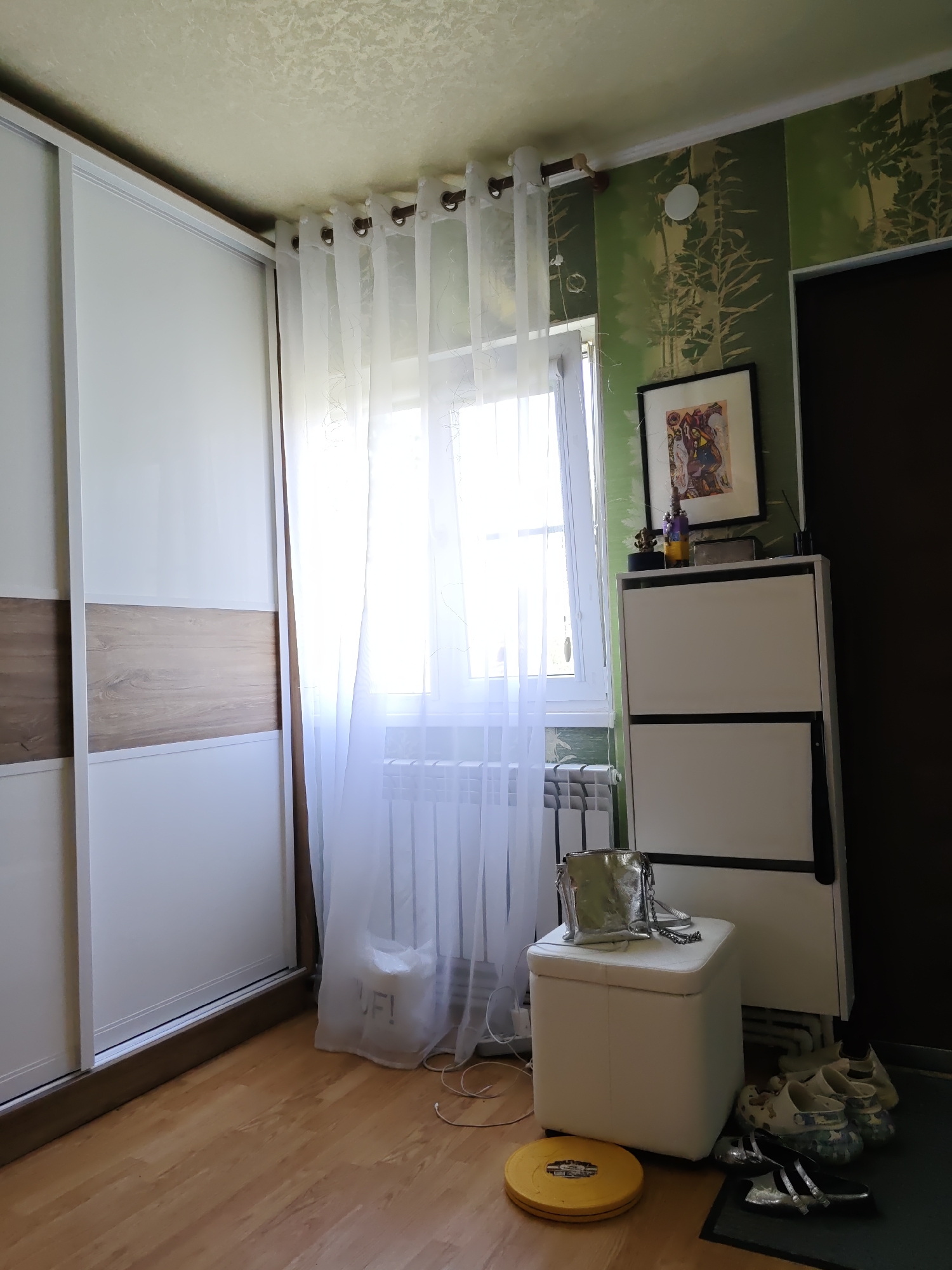 The image depicts an interior scene within an apartment. Dominating the left side of the frame is a large, modern white sliding door wardrobe with a horizontal wooden accent panel. To the right of the wardrobe, a white sheer curtain is drawn across a window, allowing bright natural light to filter through, illuminating a white radiator situated below the window. Further to the right, the wall is adorned with green wallpaper featuring a nature-inspired pattern. On this wall, a framed picture hangs above a white shoe cabinet with two drawers. On top of the shoe cabinet, several decorative items are placed, including a purple bottle and a small statue. In the foreground, to the right of the shoe cabinet, a white storage ottoman is present, topped with a silver metallic handbag. On the floor next to the ottoman, several pairs of shoes are visible, including clogs and sandals. A yellow disc-shaped object is also on the floor to the left of the ottoman. The flooring throughout the scene is light-colored wood laminate. The overall impression is that of a tidy, lived-in residential space.