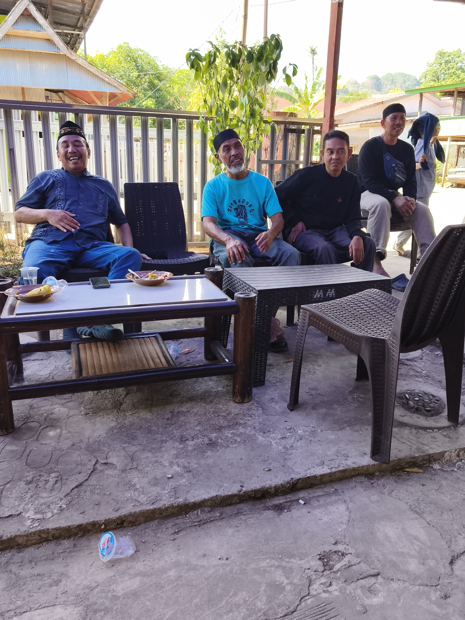 The image depicts an outdoor scene in Tonasa, Indonesia, likely a residential area or a communal gathering space. Four men are seated on chairs, with two tables in front of them. The first table, made of wood, holds two small plates with what appears to be food remnants, a mobile phone, and a clear plastic cup with liquid. The second table is a dark, modern plastic table with no items on its surface. The men are casually dressed, and some are wearing skullcaps, suggesting a cultural or religious context. The environment includes a weathered concrete floor, a metal fence, vegetation, and buildings in the background, indicating a warm, possibly tropical climate. One man on the far left is laughing heartily, while the others appear relaxed and engaged in conversation. A discarded plastic cup lies on the ground in the foreground.