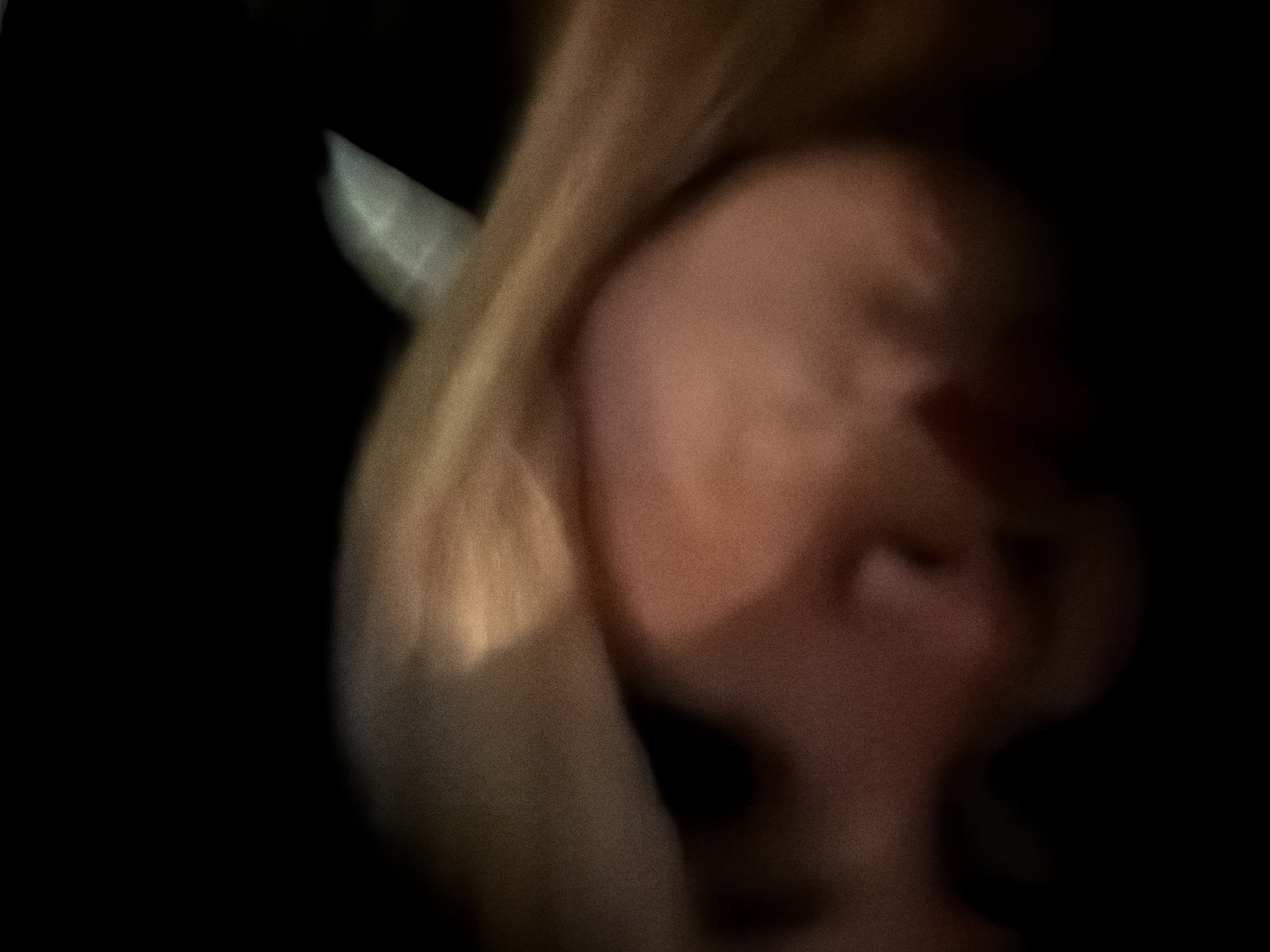 The image presents a highly blurred and indistinct close-up of a human subject, rendered in very low light conditions. The central and lower portions of the frame display a muted reddish-brown and peachy-toned expanse, consistent with human skin. Streaks of light-colored material, likely hair (blonde or light brown), traverse the upper and central areas of the frame, showing pronounced motion blur. Vague contours suggest a facial profile, including a possible nose bridge. The background is predominantly black, with a faint, geometric, light grey-white object or surface visible towards the upper right, which remains unidentifiable due to blur and low resolution. The overall scene is dark and grainy, indicative of a photograph taken in dim lighting with significant subject or camera movement. The specific location within the city of Paris, France, cannot be determined from the visual content of the image.