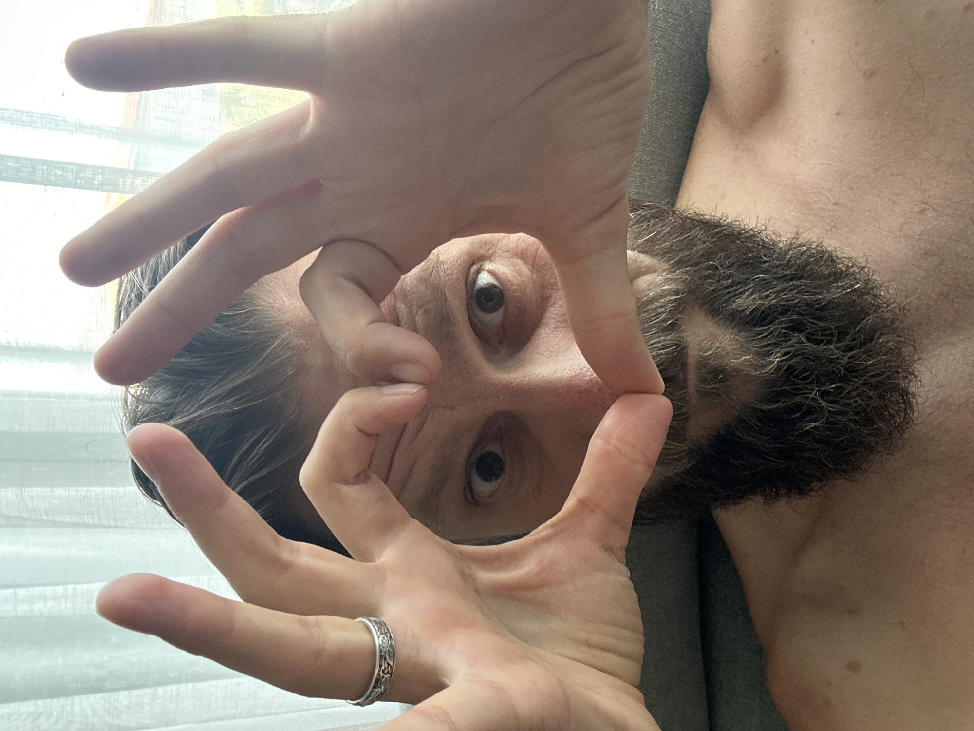 This media file features a close-up, downward-facing perspective of a person's face and upper torso. The subject has a beard and is looking directly at the camera. Their hands are positioned in front of their face, with the fingers forming a frame around one of their eyes, as if looking through a viewfinder. A silver ring with an intricate pattern is visible on one of the subject's fingers. The background suggests an indoor setting with diffused light coming from a window covered by sheer curtains. The overall mood appears playful or introspective, due to the framing of the eye and the direct gaze. The location context provided is Fethiye, Turkey. There is no visible text in the image.