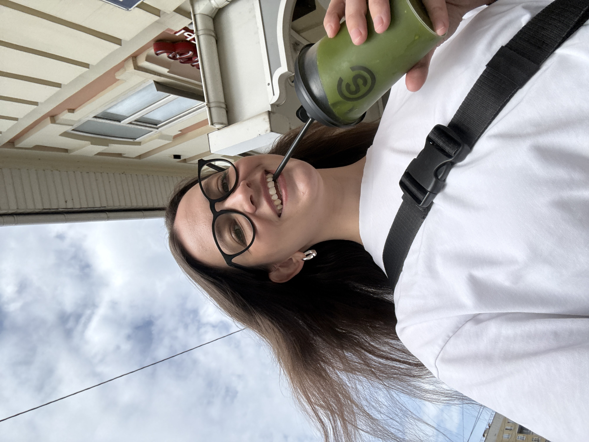 A medium shot depicts a woman in Nizhny Novgorod, Russia, holding a green smoothie in her right hand while smiling and drinking from a straw. She wears black-framed glasses and a white t-shirt with a black bag strap across her chest. The background shows architectural details of the city, including the facade of a building with windows and ornate trim, set against a cloudy sky.