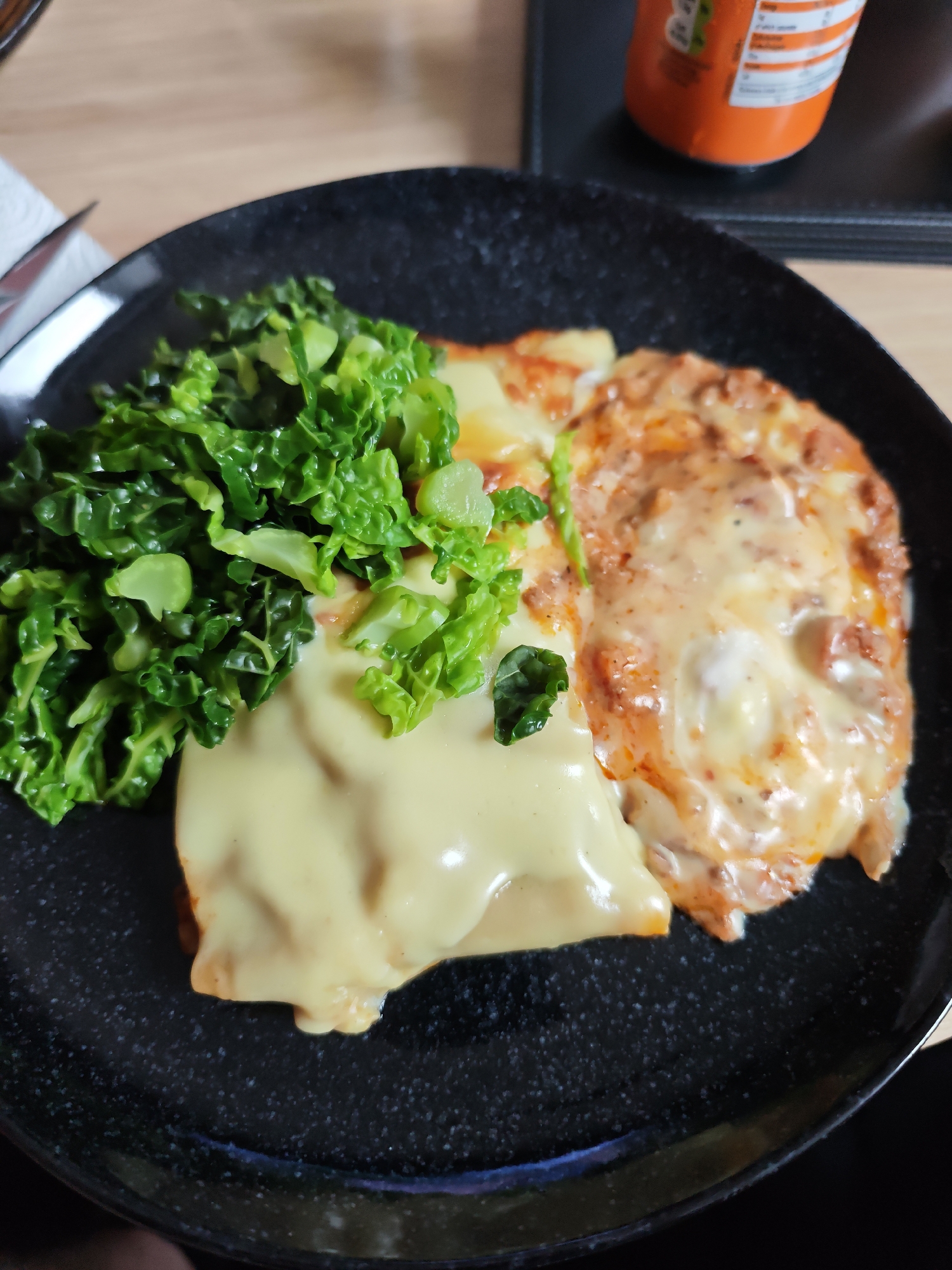 The image presents a close-up, overhead view of a prepared meal served on a speckled black plate. The plate holds three main components. To the left, a vibrant pile of green, leafy vegetables, likely kale or collard greens, appears lightly cooked. Adjoining these greens, a portion of food is covered in a smooth, creamy, pale yellow sauce, suggestive of a cheese sauce. To the right, another section of food is blanketed in a rich reddish-orange sauce, possibly a meat or tomato-based sauce, topped with melted and slightly browned cheese, resembling a baked pasta dish like lasagna.
The plate has a textured, matte black finish with subtle lighter speckles. In the upper left corner of the frame, a corner of a white paper napkin and the metallic handle of a utensil are partially visible. The meal is set on a light-colored wooden surface, likely a table. In the upper right background, part of an orange can, possibly a beverage, is visible. Some small, blurred text and numbers, including "0g" and percentages, are faintly discernible on the can's label, likely nutritional information. The scene depicts a ready-to-eat meal in an indoor setting.