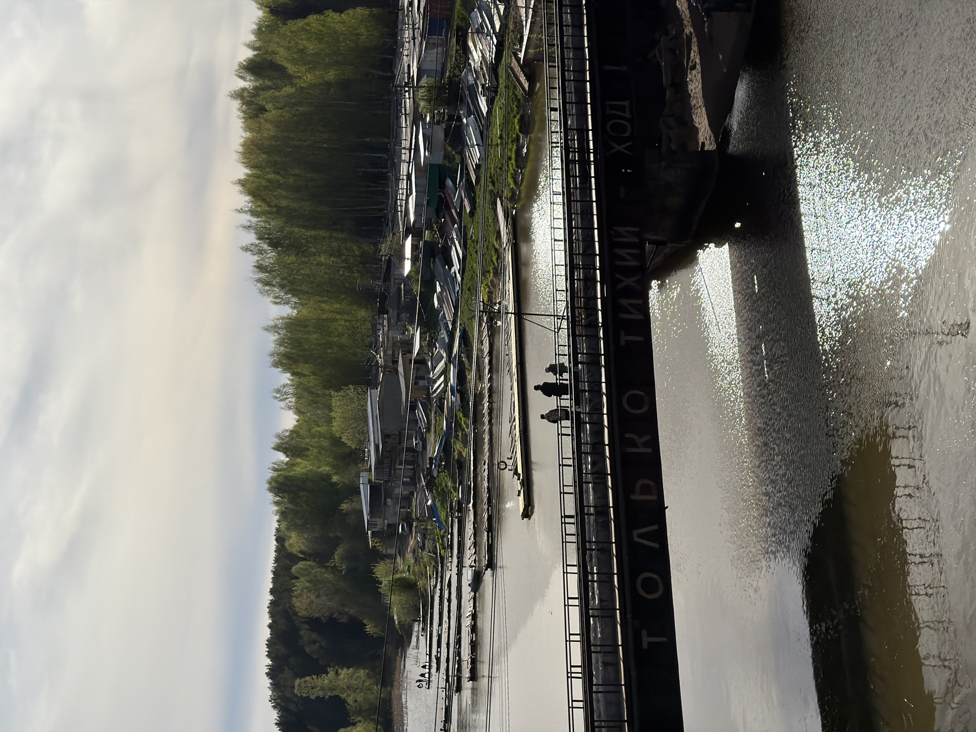 The image shows a view of the Kama River in Chaikovsky, Russia. A row of boathouses lines the riverbank, adjacent to a dense forest. A metal structure, likely part of a bridge or walkway, dominates the foreground, with Russian text visible on its side. Three figures can be seen on the walkway over the water. The river reflects sunlight, creating a shimmering effect. The sky is overcast.