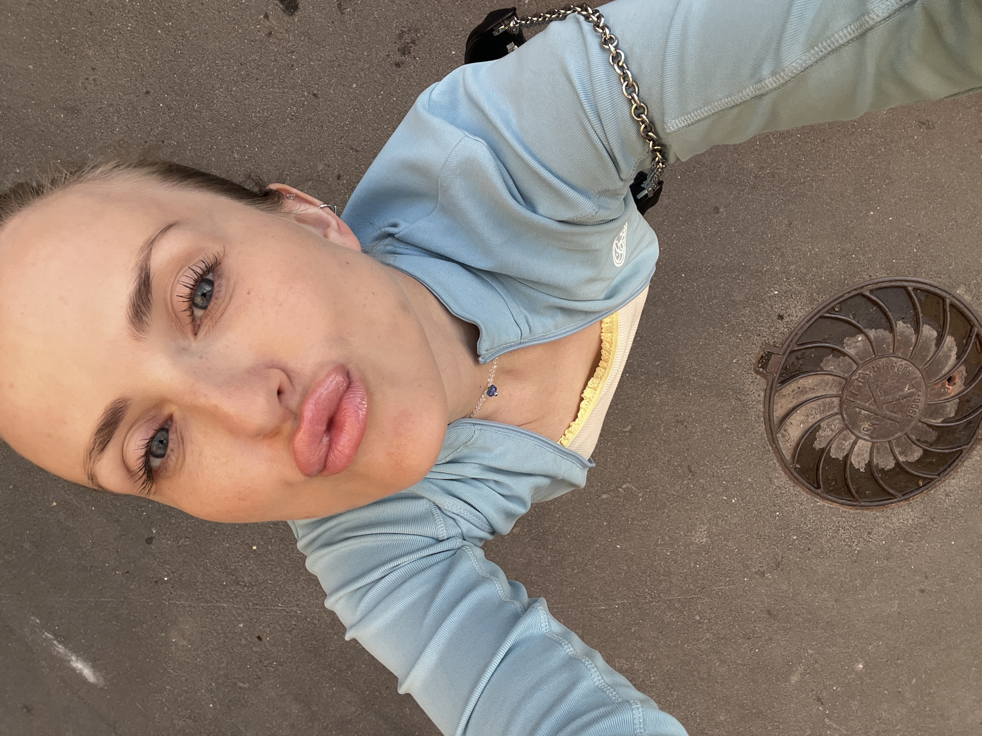 A woman with light skin and blue eyes is posing for a selfie outdoors on a paved surface. She is wearing a light blue zip-up jacket over a white top with yellow trim and a simple necklace. Her lips are pursed. In the foreground, a circular metal grate with an intricate spiral design is visible on the ground. The background appears to be an urban environment, though specific details are not clear.