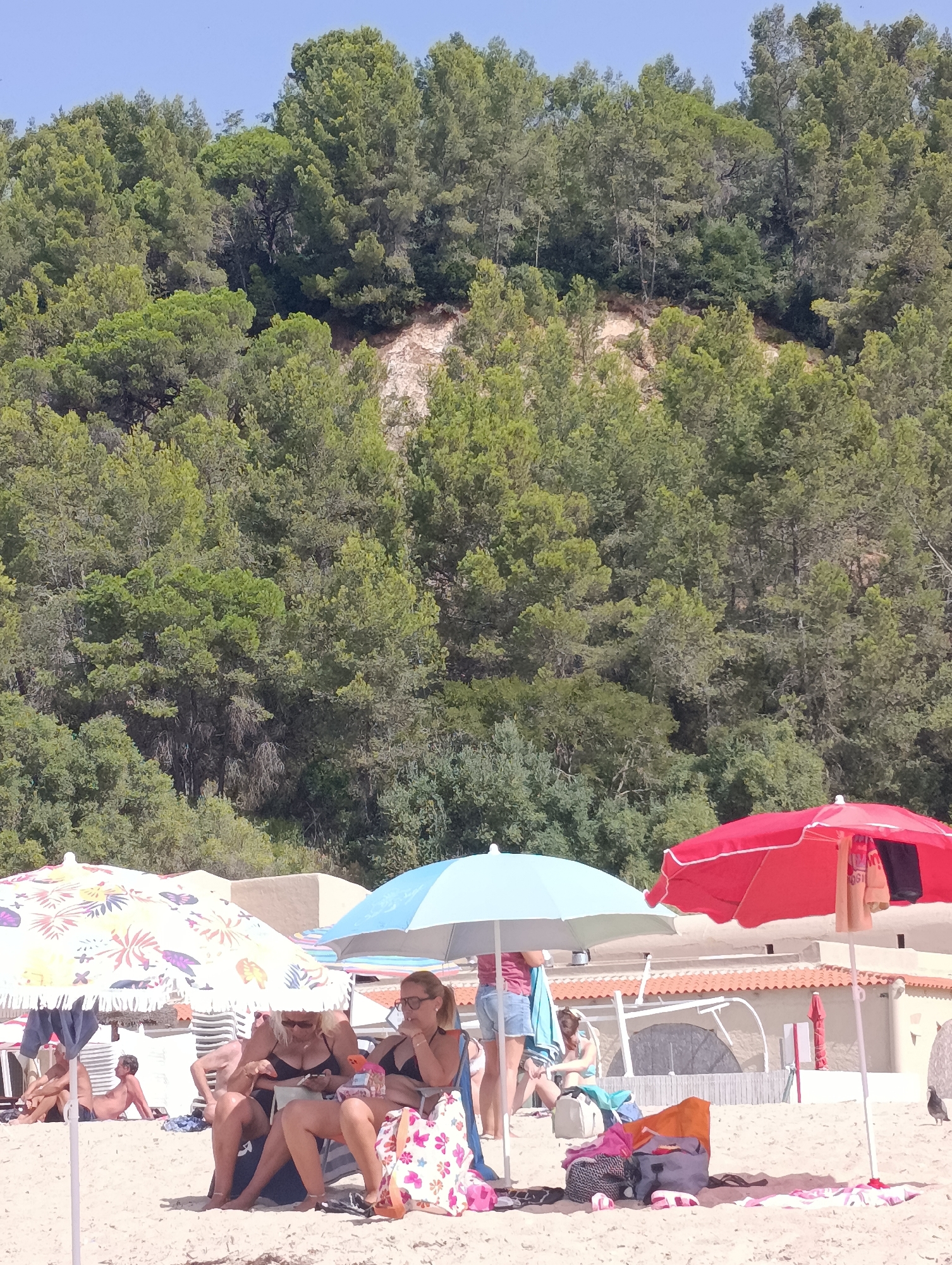 The image depicts a sunny day at a beach, likely in Setúbal, Portugal, with a densely wooded hill serving as the backdrop. Several people are present, relaxing under beach umbrellas. In the foreground, two women are seated on chairs under a colorful umbrella. One woman, with blonde hair, is looking at her phone, while the other, wearing sunglasses, is holding a cigarette and looking away. A patterned beach bag rests at her feet. To the right, a red umbrella shades another area of the beach, with some clothing draped over its pole. A light blue umbrella is positioned between the red and colorful umbrellas. Further back, a man is seen lying down on the sand, partially shaded by an umbrella. Beach chairs and other beach accessories are scattered on the sand. The background features a tan-colored building with an orange-tiled roof. The overall scene conveys a relaxed atmosphere of people enjoying a day at the beach.