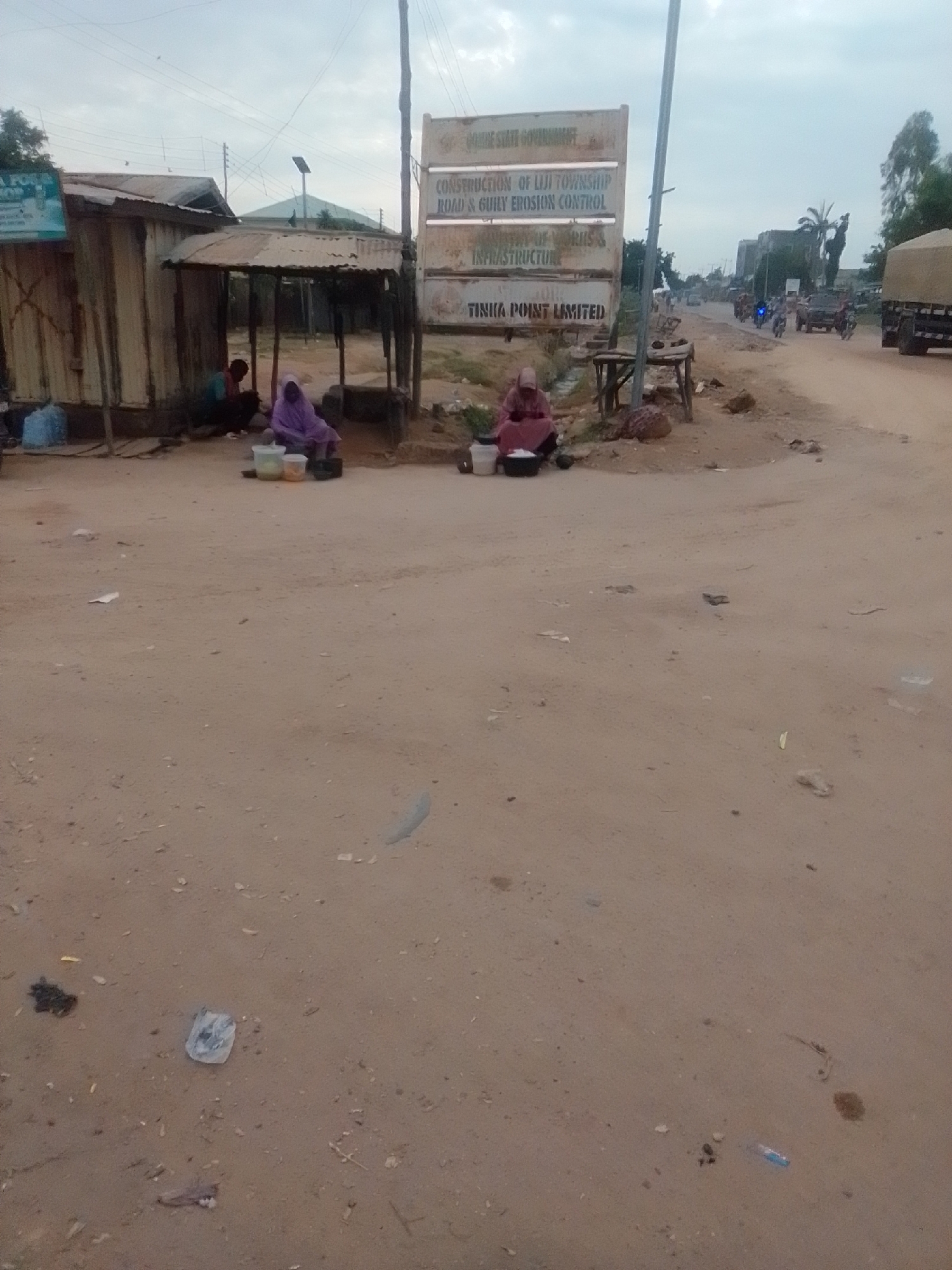 The image shows a street scene in Gombe, Nigeria, with a focus on a roadside vendor and a government information board. The ground is dusty and unpaved, typical of many areas in the city. On the left, a simple stall made of corrugated metal provides shade for two individuals seated. One person, wearing a blue and red shirt, is partially obscured, while another, dressed in a purple hijab, sits with several plastic containers in front of them, suggesting they are selling goods. The government sign, prominently displayed, lists "GOMBE STATE GOVERNMENT" at the top, followed by "CONSTRUCTION OF ILIJI TOWNSHIP ROAD & GULLY EROSION CONTROL" and "MINISTRY OF WORKS & INFRASTRUCTURE," with "TINUHA POINT LIMITED" at the bottom. Beyond the stall and sign, a road stretches into the distance, with a few motorbikes and a large truck visible, indicating ongoing traffic and commercial activity. The sky is overcast, suggesting a cloudy day.
