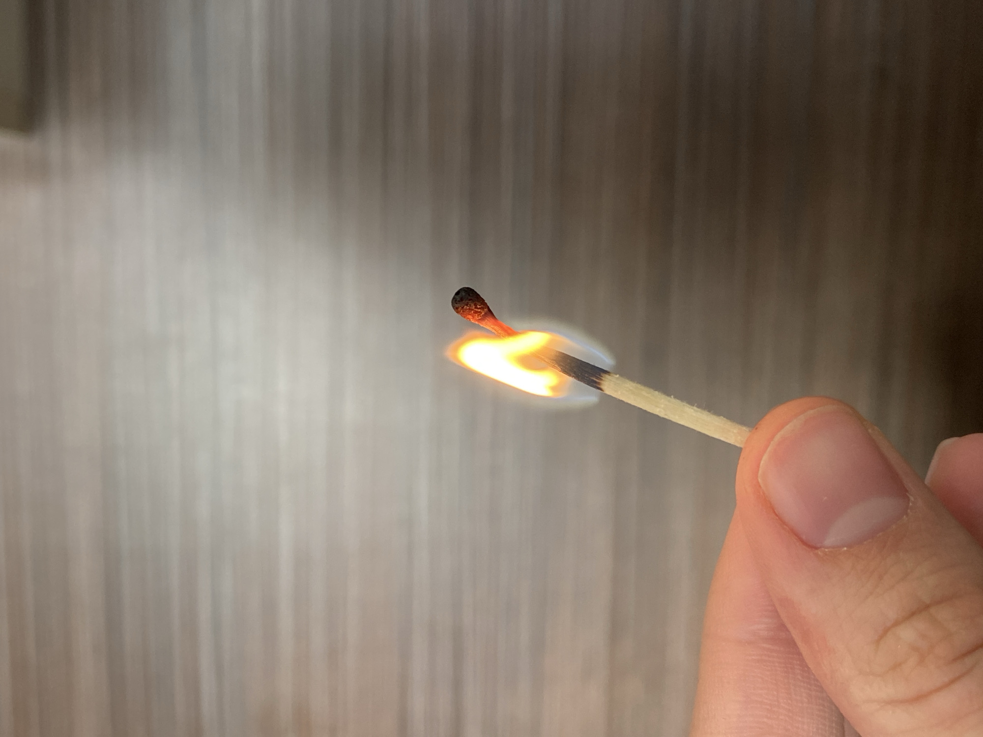 A close-up, eye-level shot features a lit matchstick held by a person's hand against a softly blurred, vertically textured background. The wooden matchstick, angled upwards from the lower right, has a vibrant flame at its tip, displaying distinct orange and blue colors. The match head is actively burning, glowing a deep reddish-orange, and the wooden shaft immediately below the flame shows signs of charring.
A human hand, likely an adult's, securely grips the matchstick with the thumb and index finger. The skin appears fair, and the fingernails are visible and well-maintained. The background is an indistinct blend of light and dark brown vertical stripes, suggesting a wooden wall or textured surface indoors. The diffused lighting illuminates the scene softly, creating no harsh shadows. There are no visible texts or specific location identifiers within the image itself, other than the provided context of Votkinsk, Russia. The scene captures the immediate aftermath of a match being struck, with the flame burning brightly.