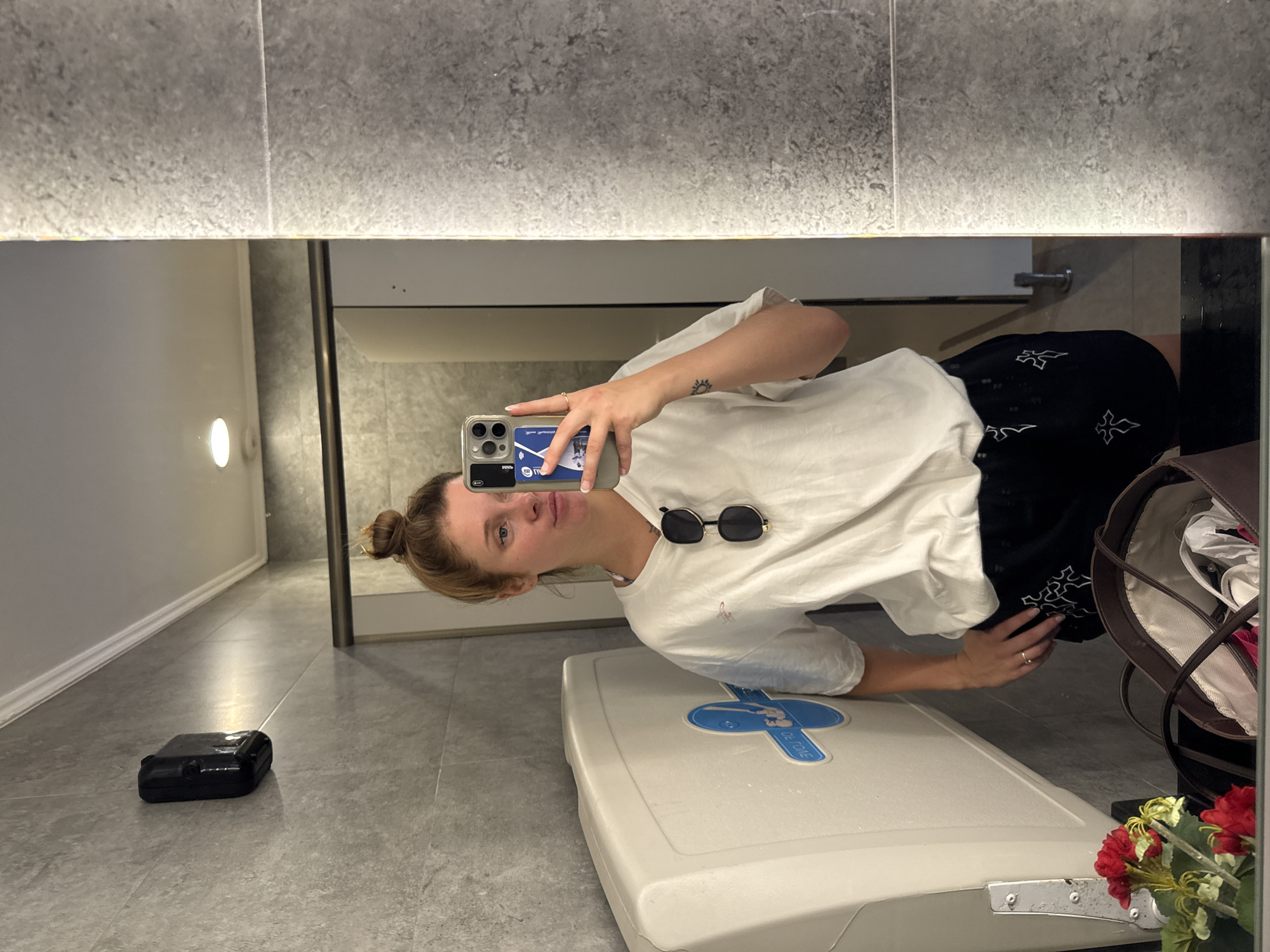 A woman with blonde hair pulled into a bun is taking a selfie in a bathroom. She is wearing a white t-shirt and black shorts with a white cross pattern. She has sunglasses perched on her shirt and a ring on her left hand. She is holding a phone in her right hand, which shows a case with two cards inserted. To her left is a white baby changing station mounted on a gray tiled wall. A black dispenser is mounted on the wall above the changing station. In the foreground, reflected in the mirror, is a brown bag and a small bouquet of red and yellow flowers. The bathroom has gray tiled walls and a white ceiling with a recessed light. This appears to be a public restroom in a facility that has a baby changing station, possibly a mall, airport, or public building. There is no information in the image to suggest the location is in Aksu, Turkey.