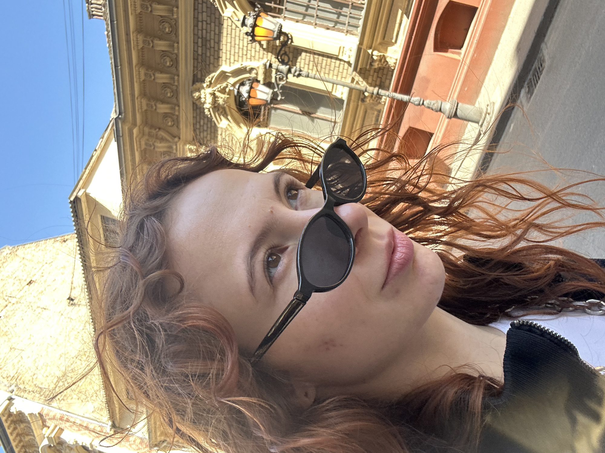 A selfie of a woman with curly red hair and sunglasses, outdoors on a sunny day. She is wearing a black jacket with a silver chain. The background includes buildings and a street lamp. This image appears to be in Novi Sad, Serbia based on the architectural style.