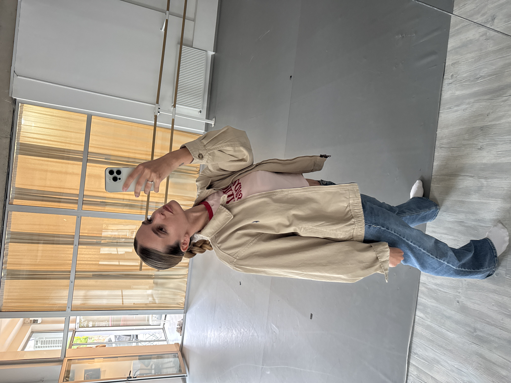 A woman stands in a room with large windows, holding a smartphone and appearing to take a selfie. The windows have white frames and tan coverings. The room's floor is a light gray color. The woman is wearing blue jeans, a light pink t-shirt, and a beige jacket. Her hair is pulled back in a braid.