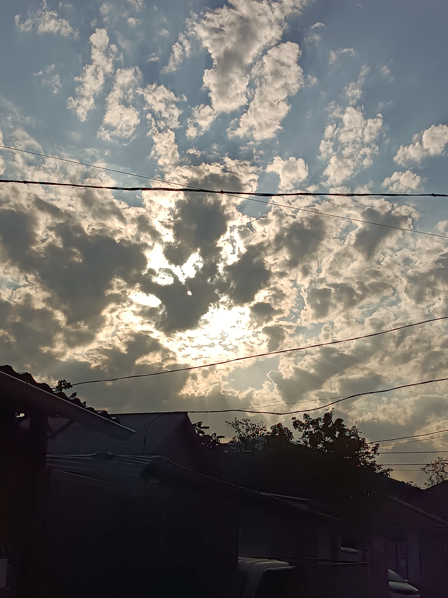 A low-angle, full outdoor shot depicts a cloudy sky with the sun peeking through. The sky transitions from a light blue at the top to a darker, grayer tone towards the horizon, filled with scattered white and gray cumulus clouds. The sun's rays are visible, creating a dramatic effect as they pierce through the cloud cover. In the foreground, the roofs of several buildings are visible, casting shadows. Power lines are strung across the sky, adding a man-made element to the natural scene. The overall impression is one of a late afternoon or early evening sky in an urban environment.