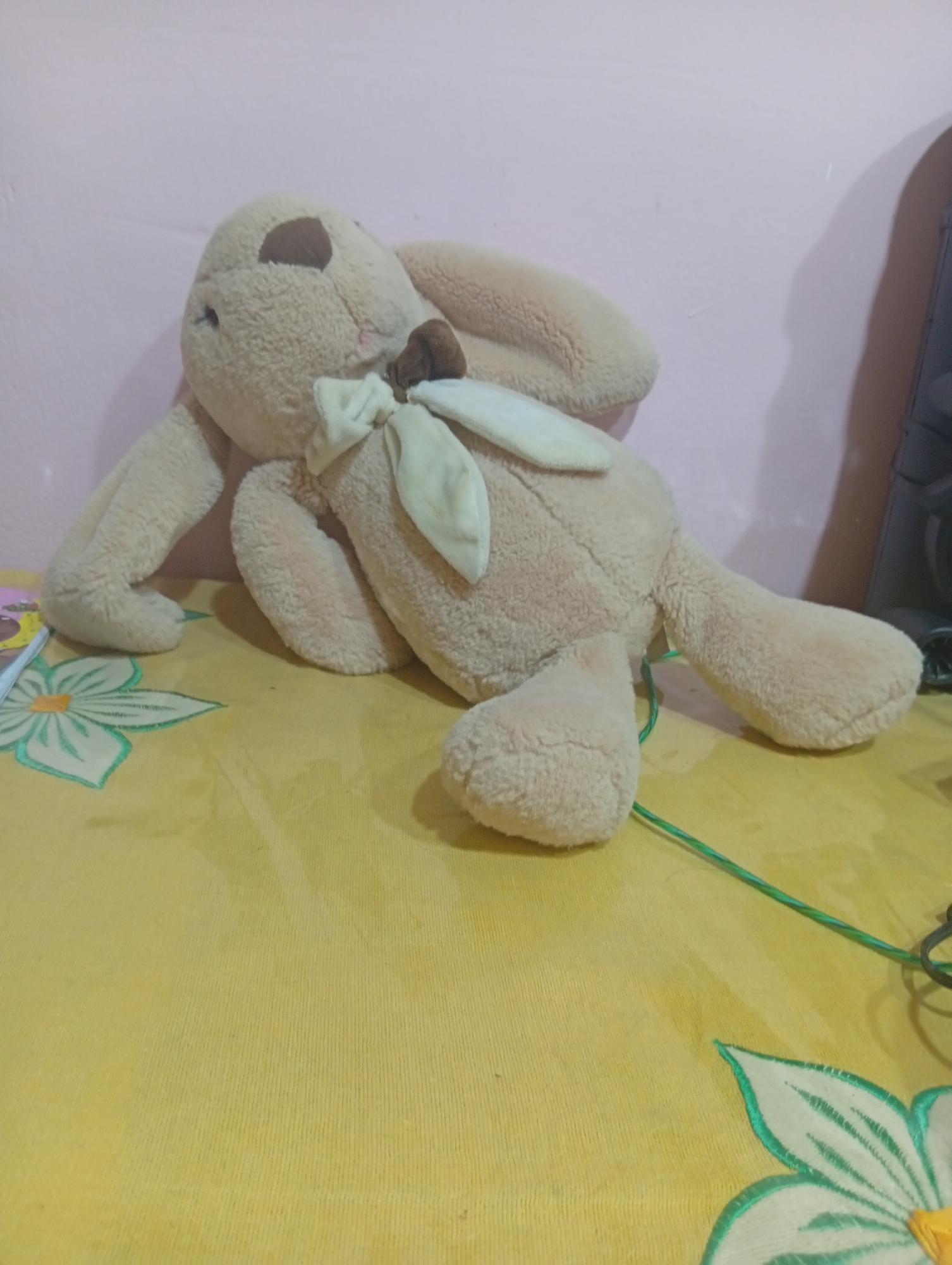 A plush bunny toy lies on a yellow tablecloth with a floral pattern. The bunny is light brown with a darker brown patch on its head and a beige bow around its neck. It is positioned on its side, with its legs extended. The background is a pink wall. The scene is indoors, likely in a residential setting in Palembang, Indonesia.