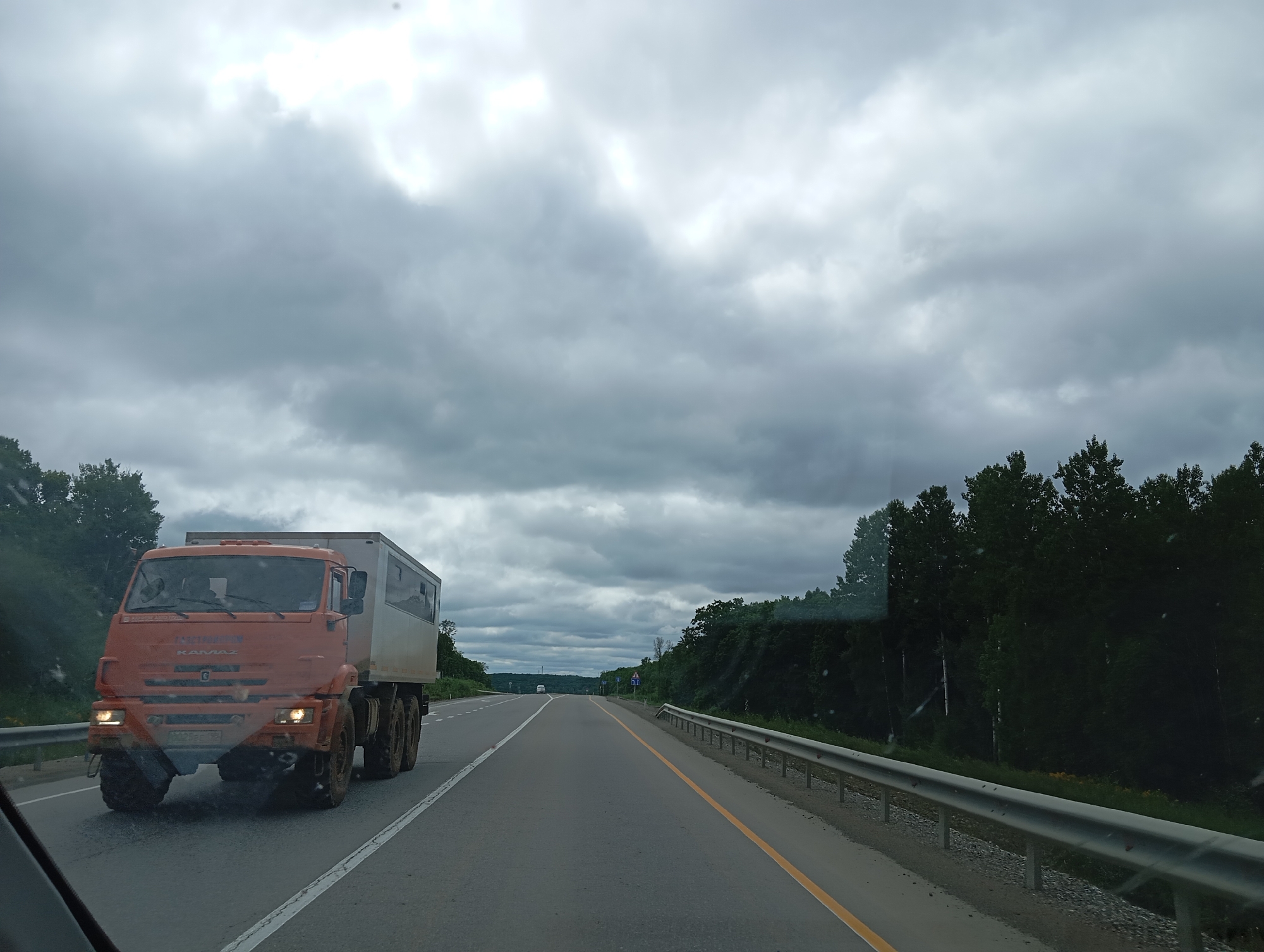 An orange Kamaz truck with a white cargo box is driving on a paved road. The truck is on the left lane, and the road has white lane markings. The truck has four wheels visible on the left side. There are trees on the right side of the road, and a metal guardrail runs along the edge of the road. The sky is overcast with gray clouds. The image appears to be taken from another vehicle on the same road, looking forward. The location is identified as Taezhnoye, Russia.