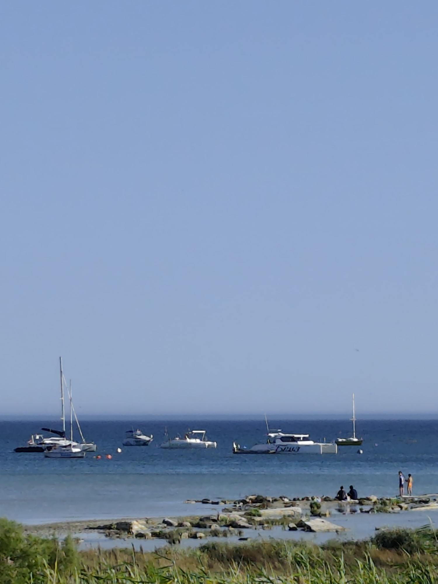 The image depicts a calm seascape with several boats anchored in the distance. The sky is a clear, pale blue. In the foreground, a rocky shoreline is visible with some vegetation growing on it. Several people are seated on the rocks, appearing to be relaxing or conversing. Further out in the water, there are at least five boats, including a sailboat with its mast and rigging clearly visible, and several motorboats. The water is a deep blue, indicating it is likely the Caspian Sea. The scene suggests a peaceful day at the beach or on the water. This scene could be located in Aktau, Kazakhstan, given its coastal location on the Caspian Sea.