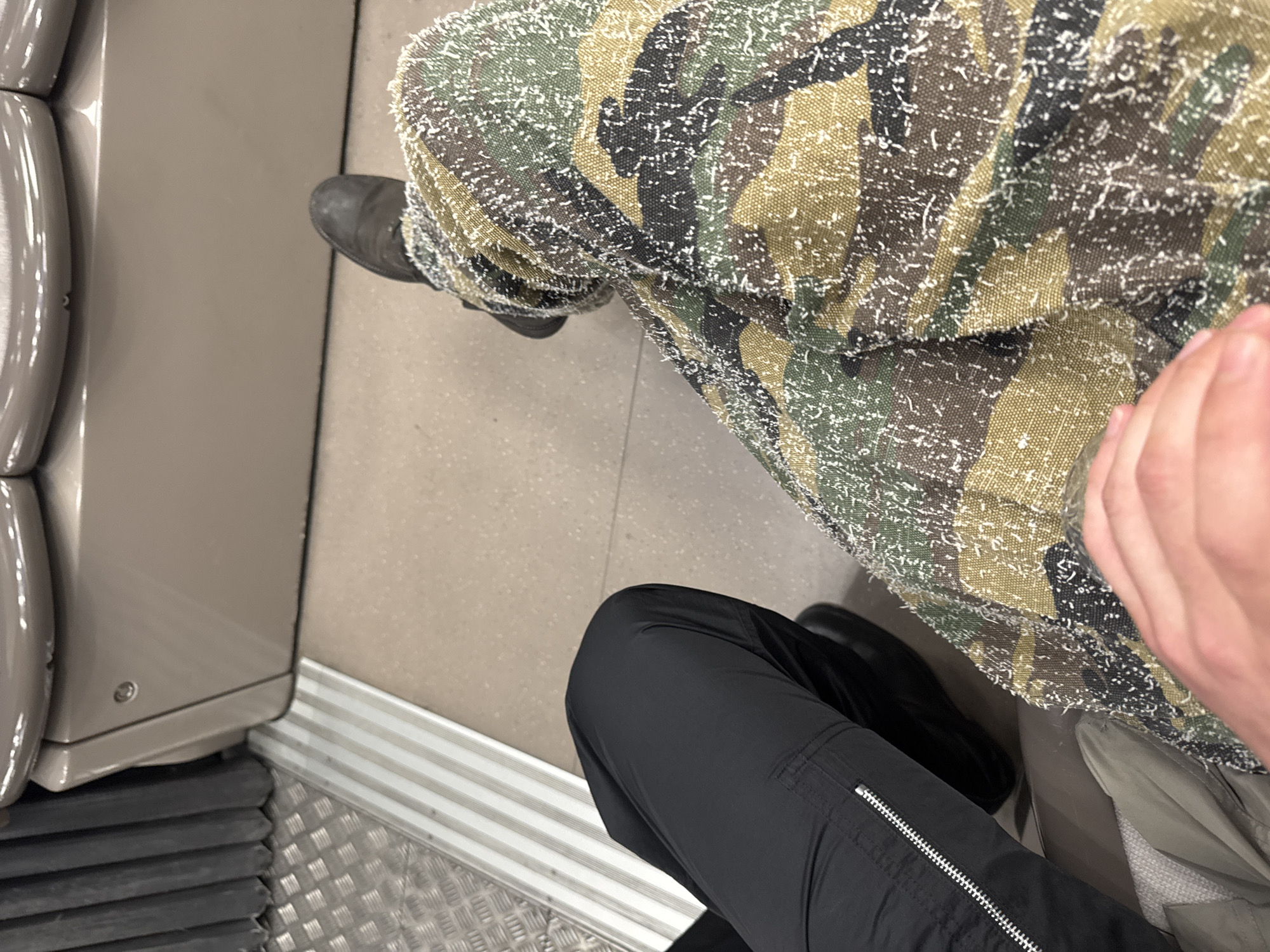 An overhead, slightly angled shot captures the legs of two individuals seated in what appears to be a public transport vehicle, consistent with a setting in Paris, France. The perspective looks down towards the floor and the people's laps.
On the right, a person wears unique camouflage patterned pants, which feature green, brown, and black tones with a distinctive white, fuzzy or thread-like texture scattered across the fabric, resembling snow or distressed material. A black boot is visible on their left foot. Their right hand rests on their knee.
To the left, another person wears plain black cargo-style pants with a silver zipper detail on a side pocket. A glimpse of a dark shoe or boot is visible near their feet. Next to them, a light-colored, possibly beige or grey, object, potentially a bag or folded clothing, is seen.
The interior environment features a smooth, light grey floor in the center, transitioning to a silver, ridged metallic strip, and then a diamond-plate textured metal section closer to the lower left. Adjacent to this, a ribbed black rubber mat is visible. On the far left, a rounded, grey/tan padded surface, likely part of a seat or interior wall of the vehicle, is present. The overall lighting is even, suggesting an indoor setting. No discernible text or specific time of day is visible.
