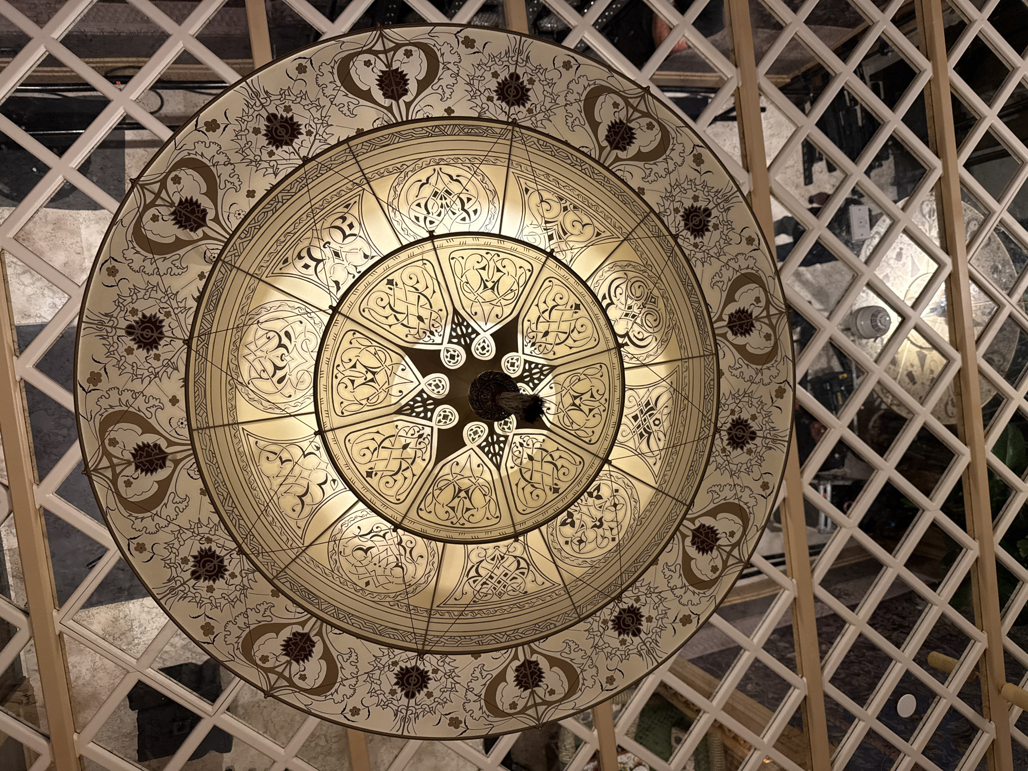 This is a low-angle, indoor shot of a large, ornate chandelier hanging from a ceiling that features a reflective, diamond-patterned grid. The chandelier is circular with multiple concentric rings of intricate design. The outermost ring is a pale cream color decorated with a repeating pattern of stylized floral motifs in brown and dark red. The next inner ring is also cream-colored, featuring a geometric pattern of interlaced lines and smaller decorative elements. The central part of the chandelier is illuminated from within, revealing a complex star-like pattern composed of flowing, dark lines against the glowing cream background. The overall aesthetic suggests a traditional or opulent setting. The reflective ceiling grid shows distorted reflections of the surroundings, including what appear to be other light fixtures, furniture, and possibly people, but they are too indistinct for clear identification. The style of the chandelier, with its detailed arabesque-like patterns, is reminiscent of Islamic or Middle Eastern design. While no specific location in Baku, Azerbaijan is identified, the design elements are consistent with architectural and decorative styles found in the city.