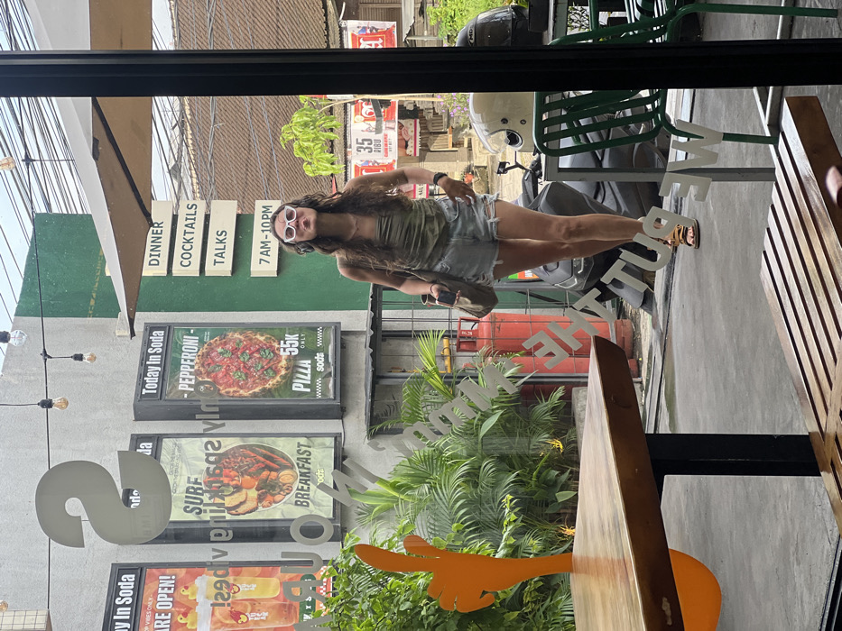 A woman with long, wavy hair and white sunglasses stands in front of a restaurant window, reflected in the glass. The window displays promotional posters for "Today In Soda" with pizza and breakfast options, along with signs for "Dinner, Cocktails, Talks." A motorcycle with a helmet sits outside, partially visible through the glass. A wooden table and green chairs are inside the restaurant. The overall image appears to be taken through a window, capturing the woman and the restaurant's exterior.