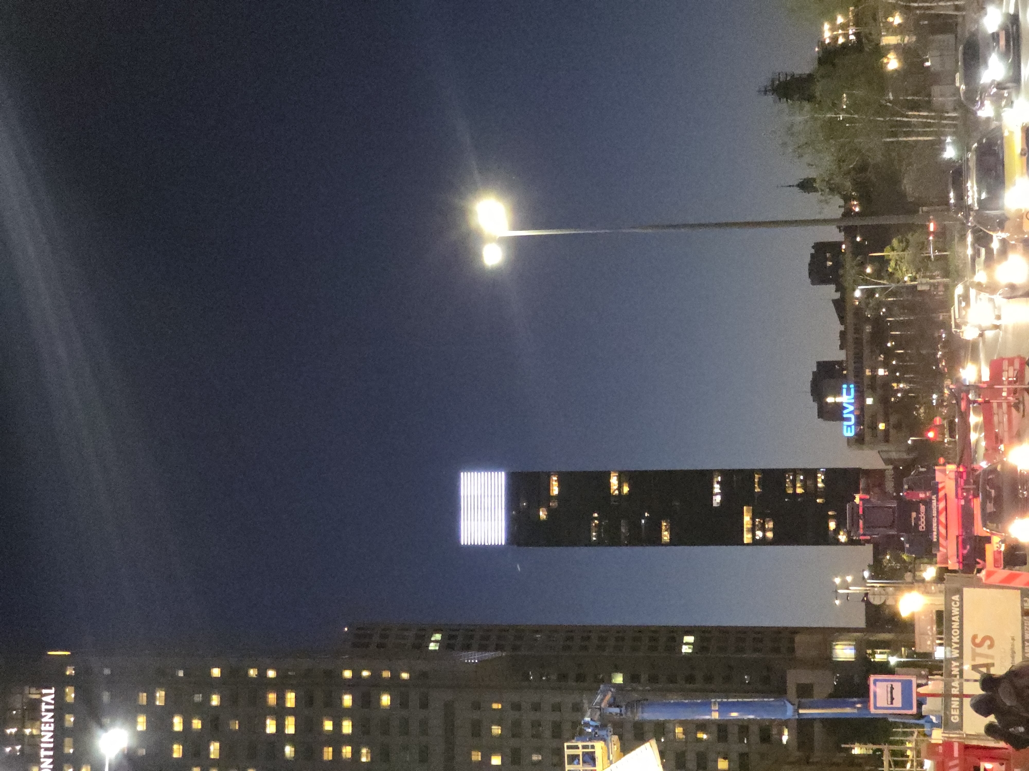 The image shows a nighttime urban scene in Warsaw, Poland. The skyline features several high-rise buildings with illuminated windows. One notable building has a vertical strip of bright white lights on its facade. Street lighting is visible, casting a glow on the surrounding area. There are several vehicles with headlights and taillights. The image appears to be taken at dusk or night, with a dark blue sky as the backdrop. Signs and other street-level details are partially visible. The image is captured from a low-angle perspective.