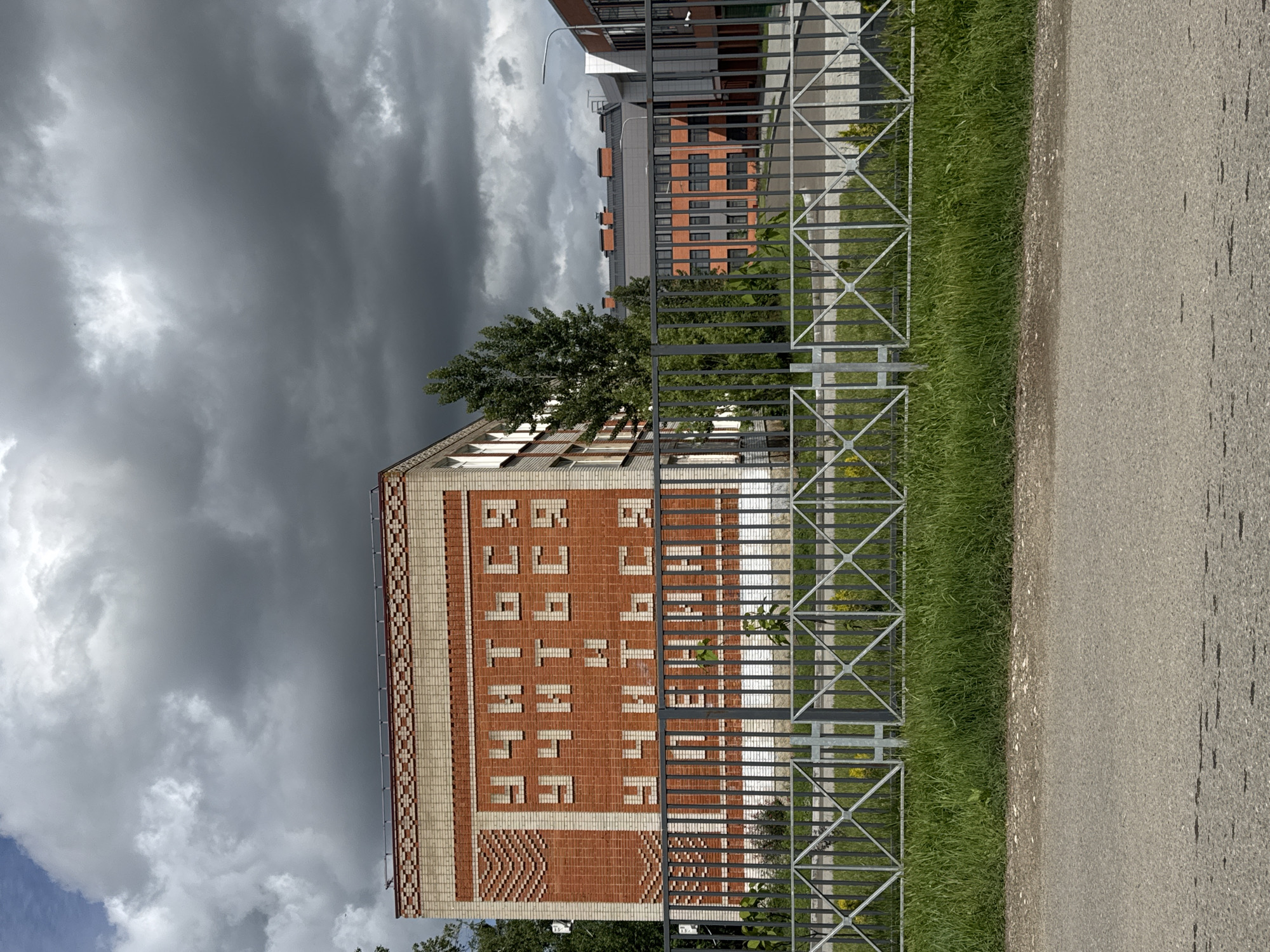 The image depicts a building in Yelizavetinskaya, Russia, near a fenced walkway. The building has a brick facade, and the words "АЛЕКСАНДРОВСКАЯ СЛОБОДА" are written vertically on the building. The sky is cloudy and overcast. The fence is made of metal. A paved path is visible next to the fence. A strip of grass grows between the fence and the path. In the background, another building is visible. A tree is between the building and the fence.