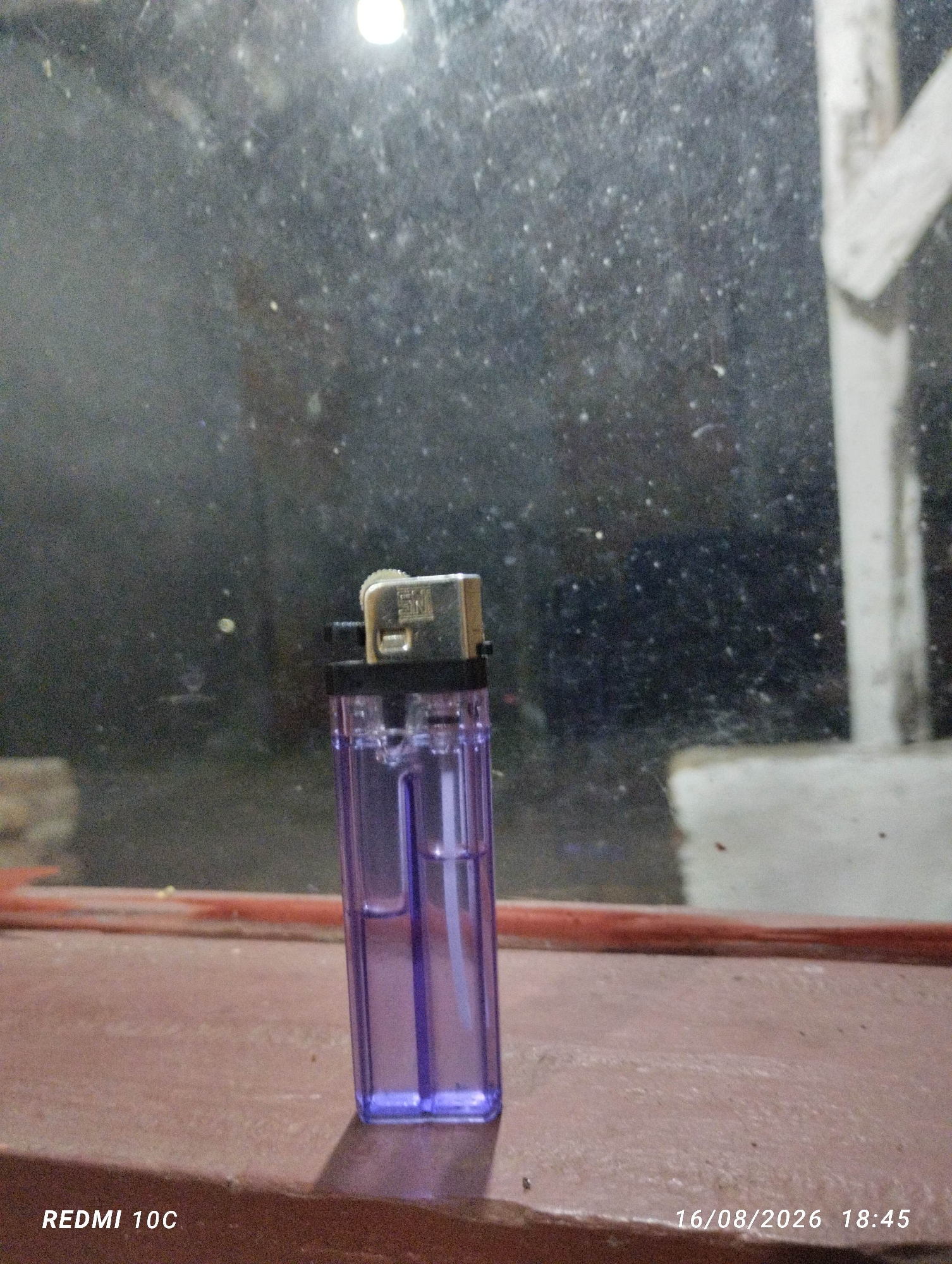 A purple plastic lighter with a metallic top is positioned upright on a reddish-brown ledge, which appears to be made of wood or concrete. The ledge is located in front of a window with a dark, speckled surface, suggesting it is either dirty or frosted. The background behind the window is blurry and indistinct, with some lighter areas that might indicate light sources or distant objects. The bottom left corner of the image displays the text "REDMI 10C," and the bottom right corner shows the date and time "16/08/2026 18:45." The overall lighting is dim, casting a slight shadow of the lighter onto the ledge.