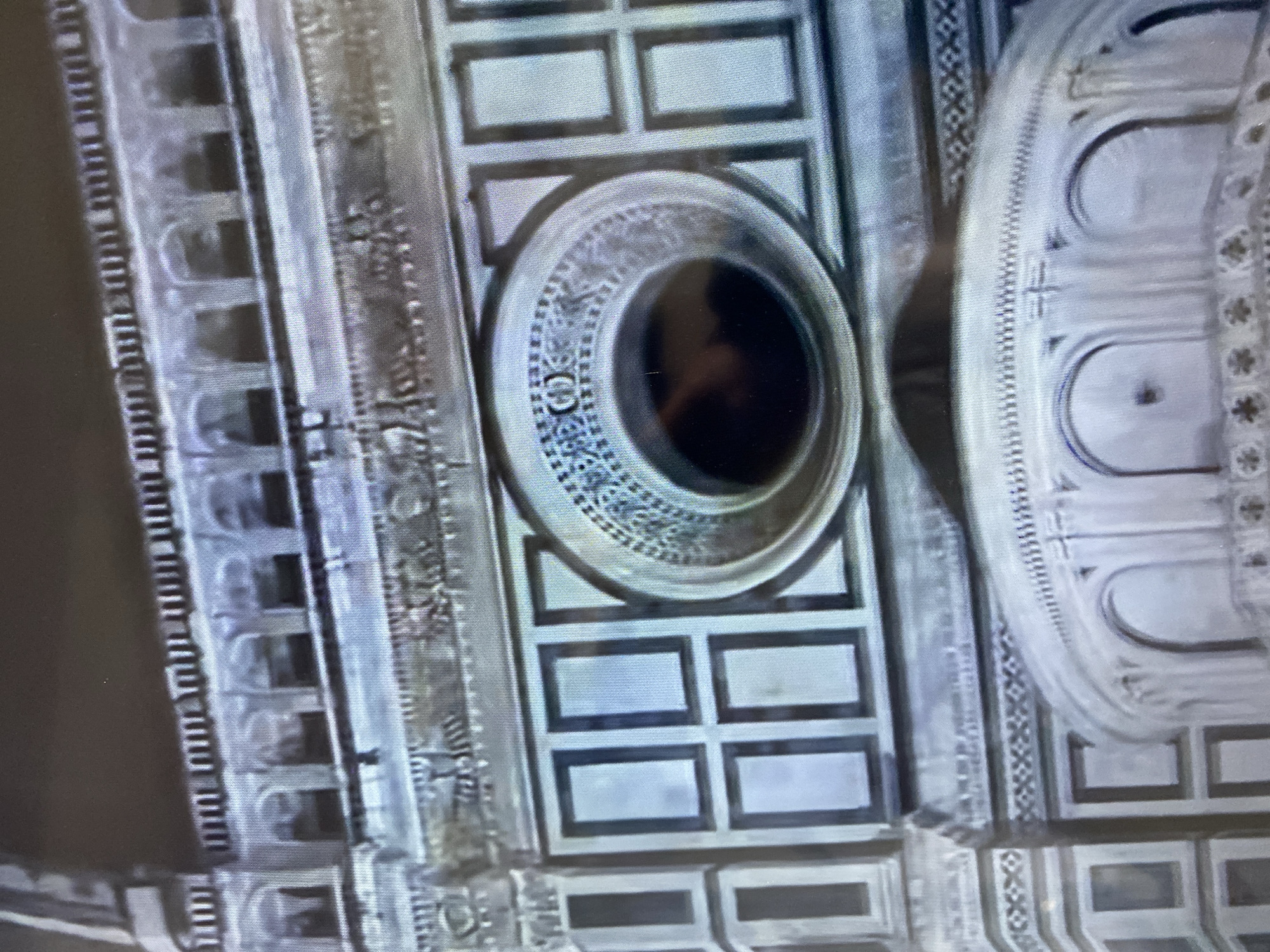 A close-up, angled forensic image displays a section of a monumental architectural facade in the city of Budva, Montenegro, characterized by ornate polychrome marble or stone construction. The scene is devoid of people, actions, or interactions.
The upper visible section features a horizontal balustrade composed of numerous small, regularly spaced arched openings, each with a dark interior, supported by slender pilasters. Above this, a simpler, darker balustrade-like structure is present. A vertical structural element is partially visible on the far left.
Below the upper balustrade, a prominent horizontal frieze traverses the facade. This frieze contains intricate bas-relief carvings depicting repeating motifs, including what appear to be stylized quadruped animals with raised tails and other symbolic figures.
The central focal point is a large, circular window, an oculus, surrounded by multiple concentric rings of elaborate ornamentation. The innermost ring is a smooth, dark circular opening, reflecting a faint, diffuse light source near its bottom-center. This is encircled by a band of regularly spaced bead-and-reel or dotted patterns. The outermost ring of the oculus features a wider band of intricate geometric designs, possibly interlocked circles or stylized floral motifs. The oculus is framed by a broad, flat dark border.
Flanking the oculus are sections of the facade adorned with a grid of light-colored, rectangular panels, each precisely framed by dark borders. Two rows of these panels are visible to the left and right of the circular window.
The lower portion of the image reveals a curved architectural element, suggesting a dome, apse, or rounded projection. This curved surface is decorated with an upper frieze exhibiting a continuous zigzag or dentil pattern. Below this, a series of evenly spaced, small arched openings or niches are visible, each containing dark interiors. Beneath these arches, another horizontal frieze displays repeating star-shaped or quatrefoil motifs.
The lighting suggests dim conditions, likely nighttime illumination, with colors muted to predominantly shades of white, light gray, and black. The image exhibits digital softness consistent with a photograph taken in low light.