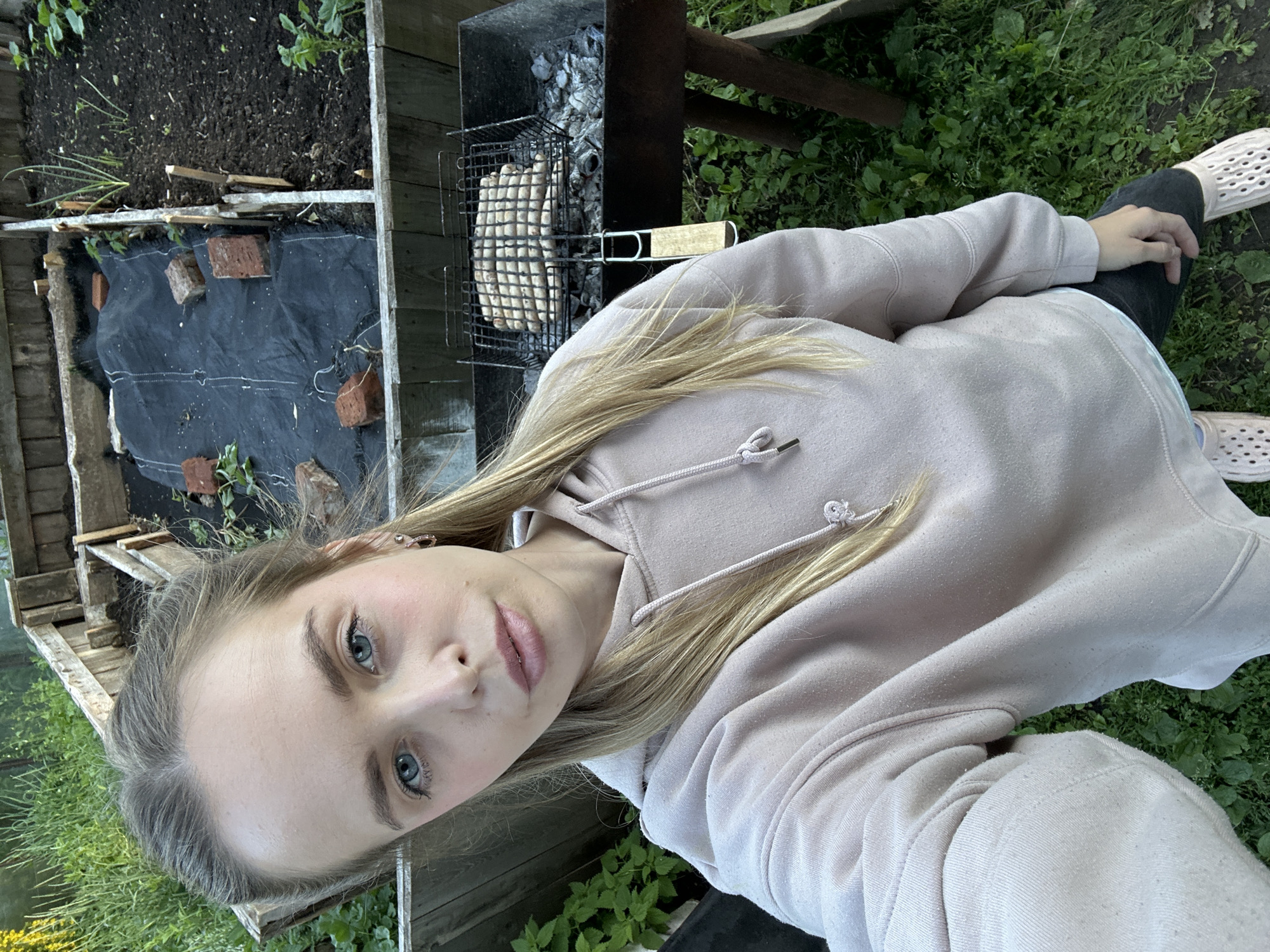 A young woman with blonde hair and blue eyes is taking a selfie outdoors. She is wearing a light pink hooded sweatshirt and dark pants, with light-colored perforated shoes. She is standing in what appears to be a backyard or garden area, with raised garden beds and greenery visible in the background. A barbecue grill with sausages on it is positioned in front of her. The environment suggests a rural or suburban setting. The location is identified as Azinsky, Russia.