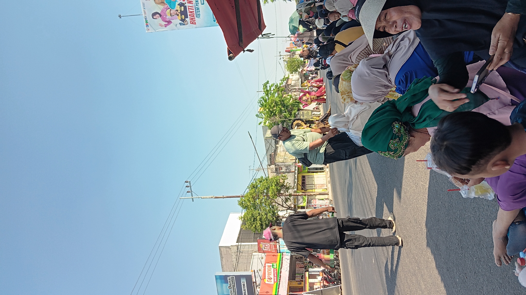 In the city of Warujayeng, Indonesia, a street scene unfolds with a group of people gathered along the roadside. The sky is a clear, bright blue. In the background, a utility pole with several wires stretches across the frame, and behind it, a billboard advertising "BHAKTI NUSANTARA SISTEM PEMILIK" is visible. Various buildings and shops line the street, including one with a sign for "GEPREKIN". A motorcycle is parked on the side of the road.
In the foreground, several individuals are seated on the asphalt, some holding drinks in plastic cups with straws. A woman in a black hijab and a wide-brimmed hat is looking towards the right, her mouth open as if speaking, and holding a smartphone. To her left, a person in a green headscarf and a pink garment is also seated. To the left of these seated individuals, two men are standing. One man, wearing a black t-shirt and black pants, has a pink cap on and is facing away from the camera. Another man in a light green t-shirt and black cargo pants, wearing a dark cap, is facing downwards. Further down the street, a procession or performance is taking place, with individuals in colorful attire, possibly traditional Indonesian costumes, walking and interacting with the crowd. The overall impression is of a public gathering or event taking place outdoors on a sunny day.