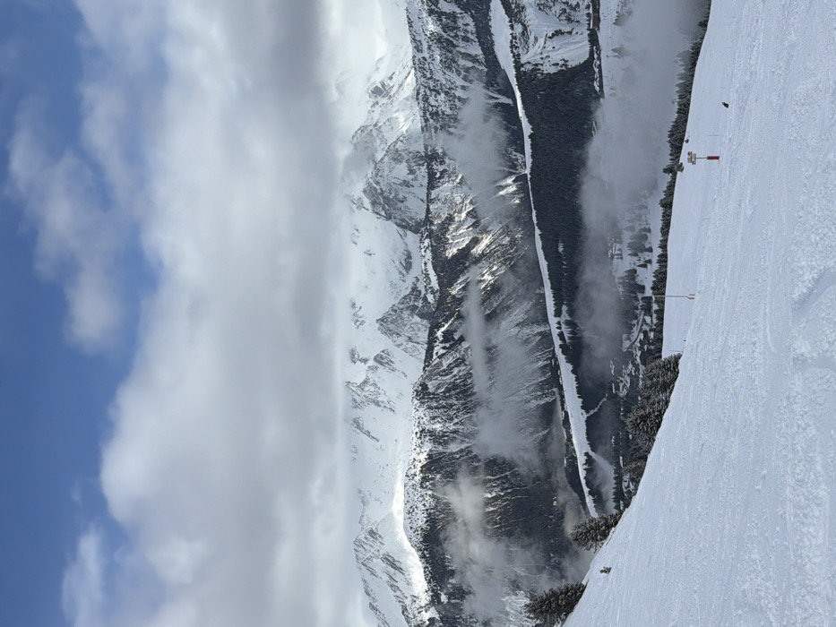 The image displays a vast snow-covered alpine landscape, identified as a ski area in Courchevel, France. In the foreground, a wide, groomed ski slope descends from left to right, covered in fresh snow with faint ski tracks. Several snow-laden evergreen trees are positioned on the upper left side of this slope. Toward the lower right, a single individual, dressed in dark winter clothing, is observed sitting or kneeling on the snow. Two ski markers are visible further down the slope: a red pole marker and a brown directional sign.
The midground reveals a deep valley partially enveloped by low-lying clouds or fog. Below the cloud line, scattered chalets and other buildings indicative of a mountain settlement are visible amongst dense evergreen forests that cover the valley slopes.
In the background, towering snow-capped mountain ranges extend across the horizon under a partially cloudy sky. Patches of sunlight illuminate certain peaks, while others are partially obscured by thick, white and gray clouds that transition into a clear blue sky overhead.