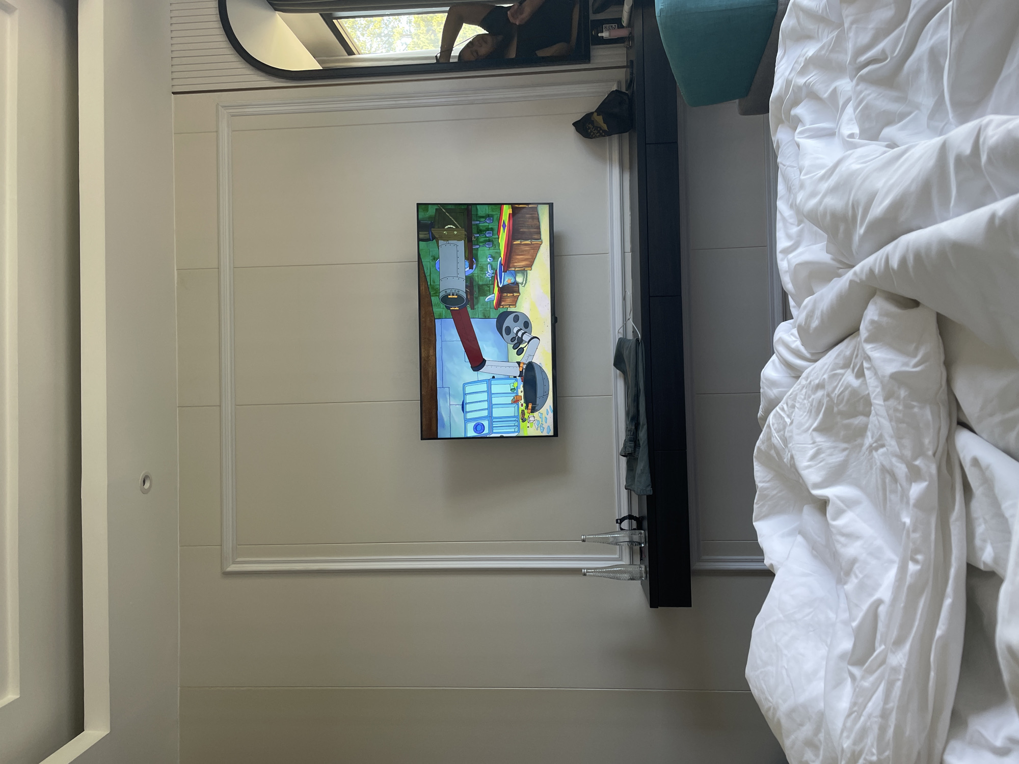 This image captures an interior scene, likely a hotel room or bedroom, in Kayuambon, Indonesia, viewed from a low perspective on a bed. The foreground consists of rumpled white bedding and a white duvet, occupying the lower third of the frame. Above the bed, a dark-colored floating console with multiple drawers is mounted against a light beige paneled wall featuring decorative white molding. On the console, from left to right, are two empty clear glass bottles, a pair of dark-framed eyeglasses, a folded garment resembling dark green or blue fabric, and a dark bag or pouch.
Centered on the wall above the console is a flat-screen television displaying an animated scene, specifically from SpongeBob SquarePants, showing a contraption with pipes, a large red arm, and various objects in a laboratory-like setting. To the right of the television, a tall, arched mirror is mounted vertically. Reflected in the mirror is a person, presumably the photographer, with dark hair and wearing a dark top, looking into the mirror. The reflection also reveals bright sunlight and green foliage outside a window, suggesting a natural outdoor view. The walls are uniformly paneled in a light beige tone with vertical and horizontal white molding accents. The ceiling features a white tray design with a single recessed light fixture visible in the upper center. A portion of a teal or turquoise upholstered chair or ottoman is visible at the far right edge of the frame.